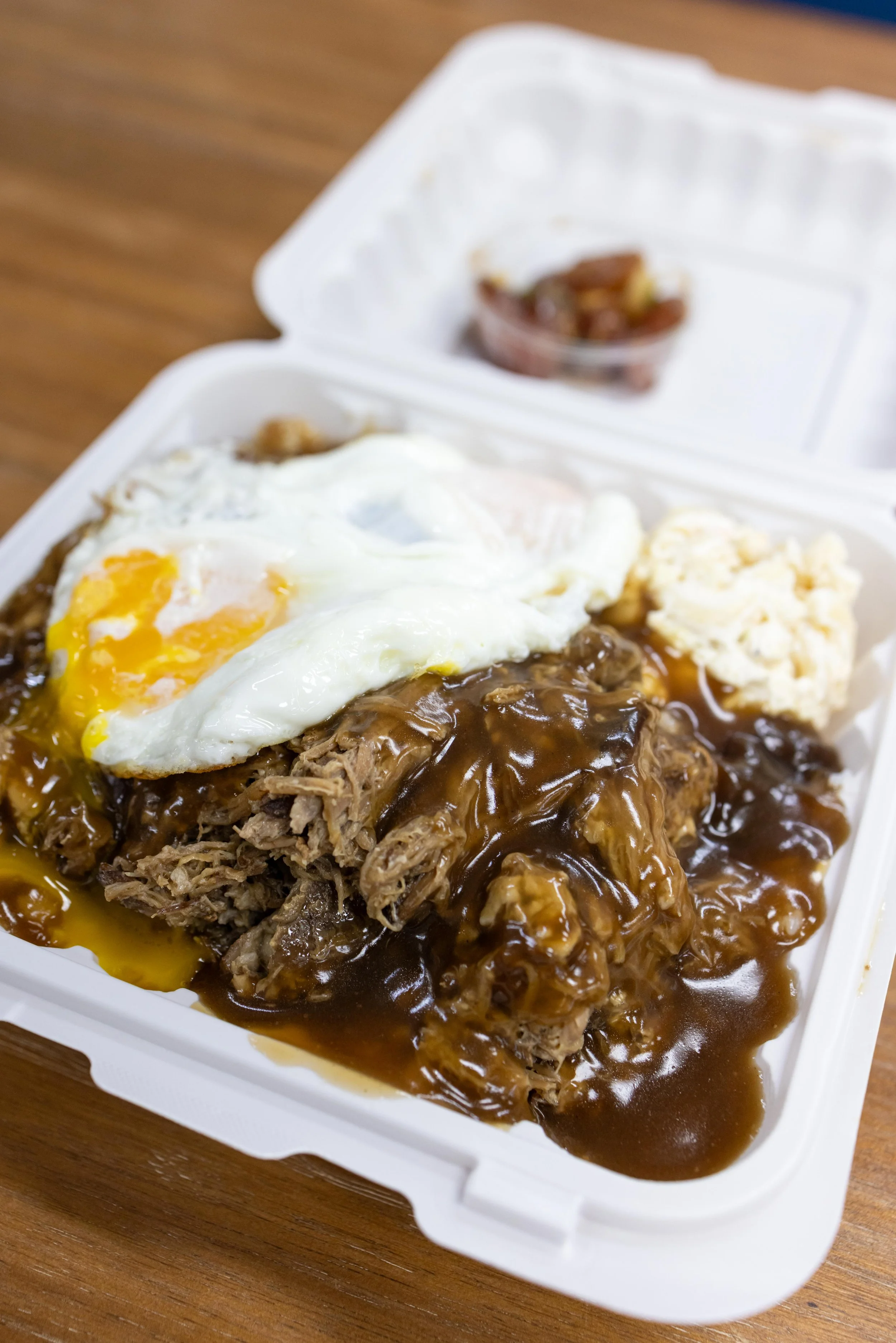 Close-up of a meatloaf with brown gravy, a fried egg on top, mashed potatoes, and a small container of diced onions in a takeout container on a wooden table.