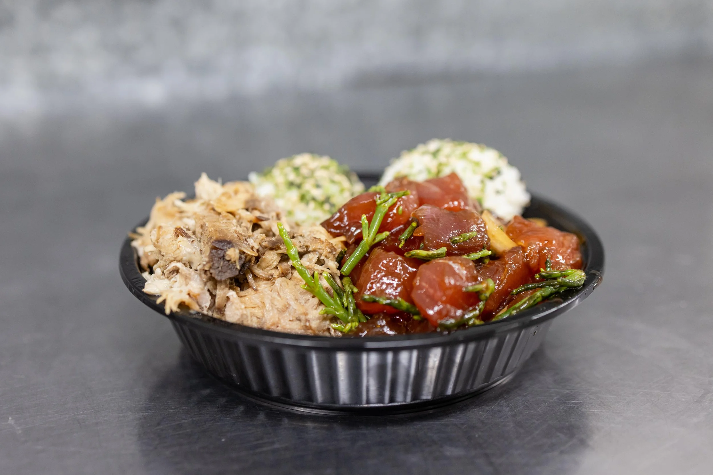 Black container filled with shredded meat, braised beef stew with green vegetables, and two scoops of potato salad garnished with herbs.
