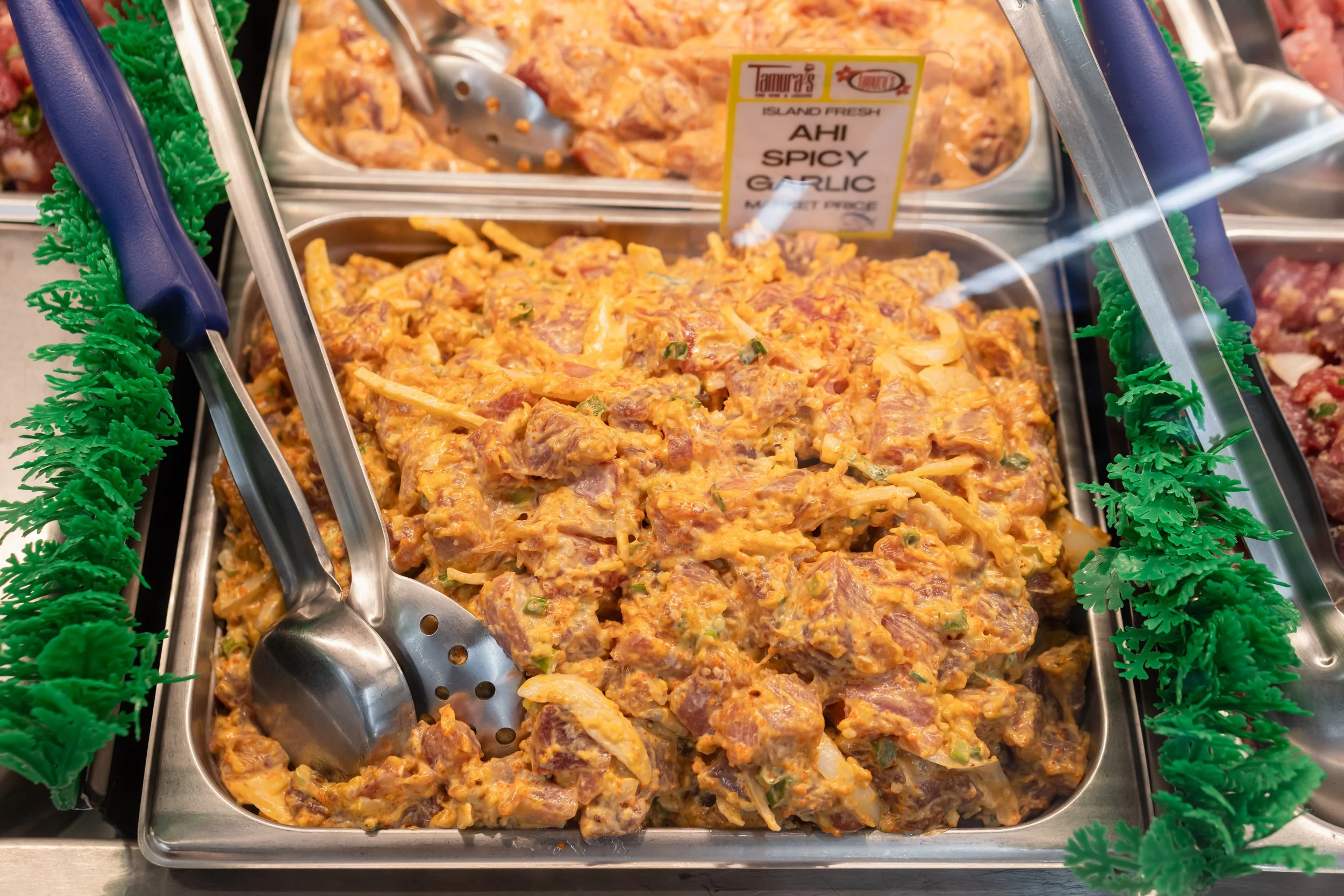 A metal tray of Asian spicy garlic marinated meat at a buffet, with a pair of tongs resting on top, decorated with green plastic greenery along the edges.