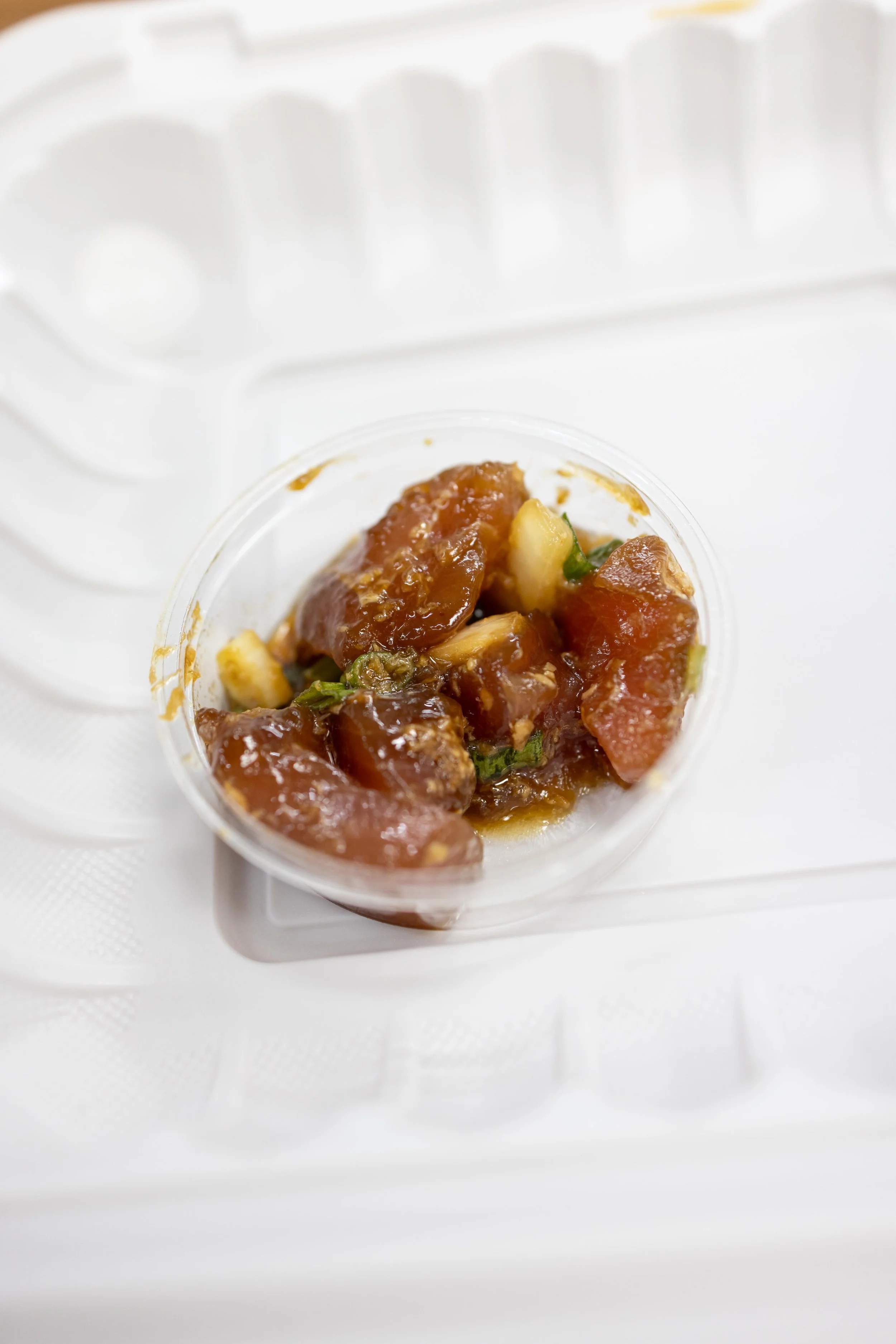 Small container of chopped raw fish or seafood with sauce and green garnish, placed on a white surface.