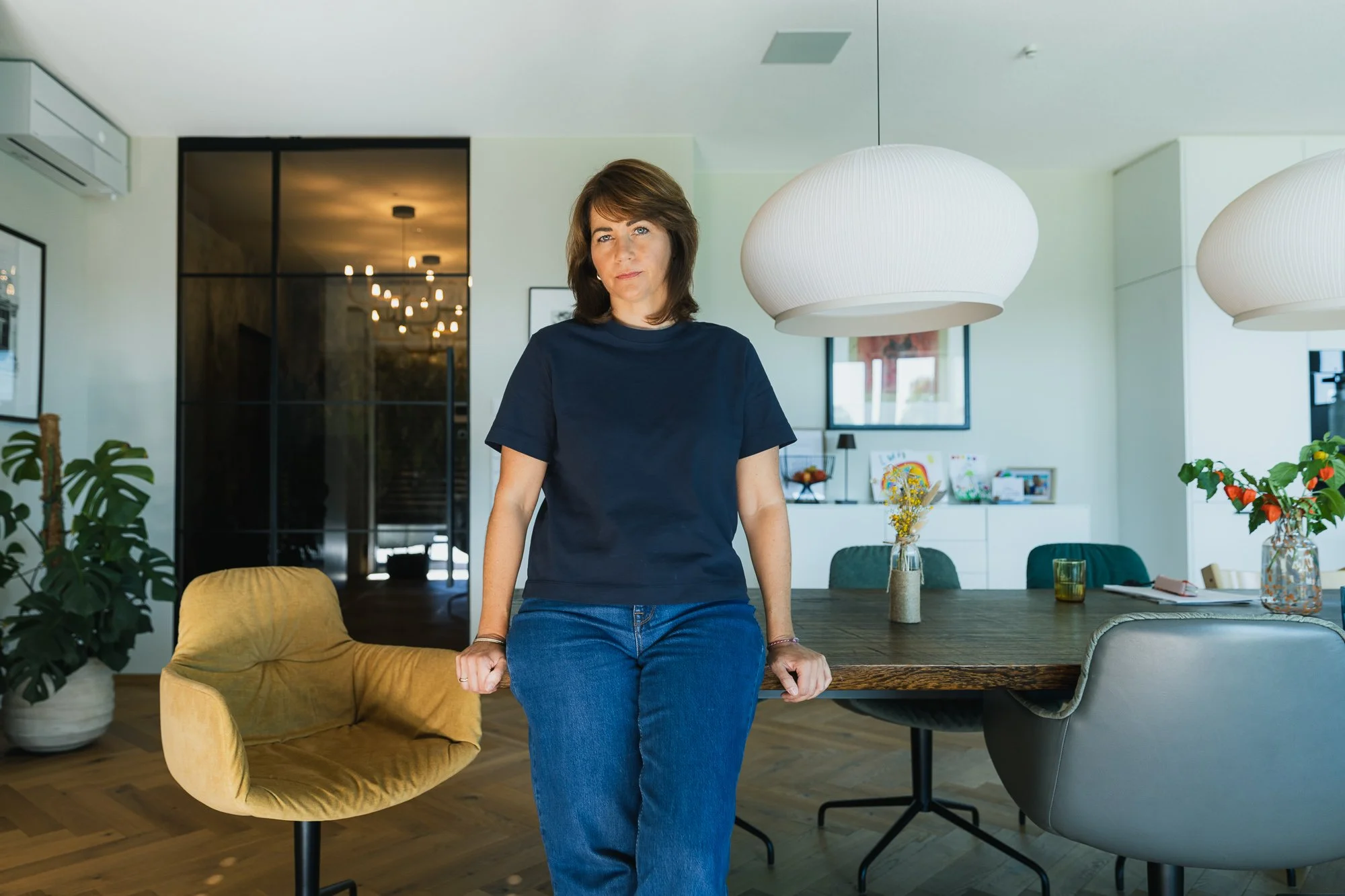 Eine Frau in einem dunkelblauen T-Shirt steht in einem modernen, hellen Esszimmer mit grünen Stühlen und hellen Lampen. Im Hintergrund sind Tische, Pflanzen und Fenster sichtbar.