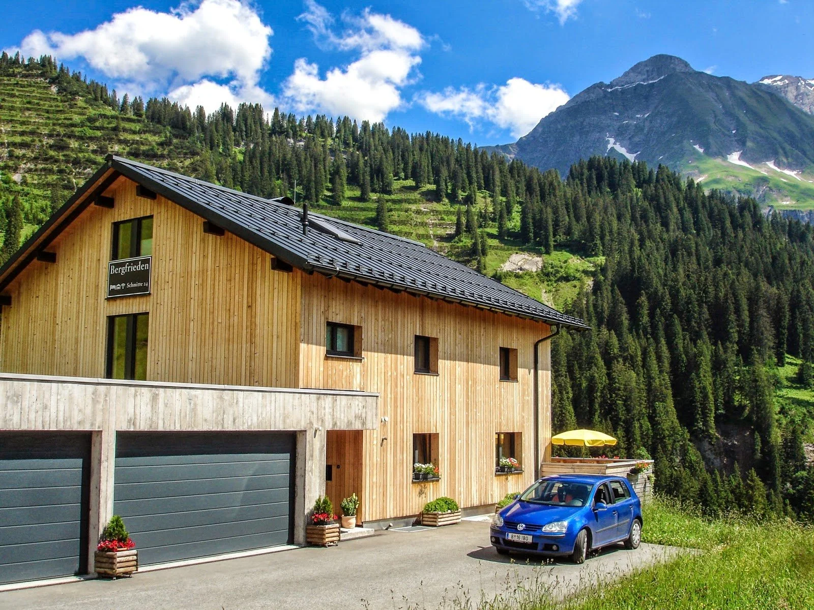 Ein mehrstöckiges Holzhaus mit zwei Garagentüren, kleinen Fenstern und einem blauen Auto davor, vor einer bergigen Landschaft mit Nadelwäldern und einem hohen Berg im Hintergrund.