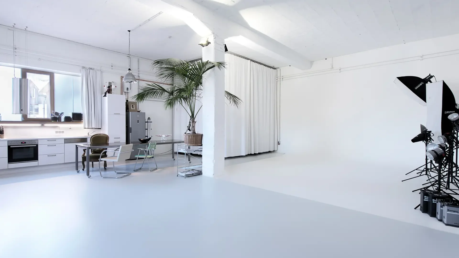 Ein weißer Raum mit einer modernen Küchenzeile, einem großen Pflanzen in einem Korb, einem weißen Vorhang, und Fotostudio-Ausrüstung wie Beleuchtung und Kameraständer.