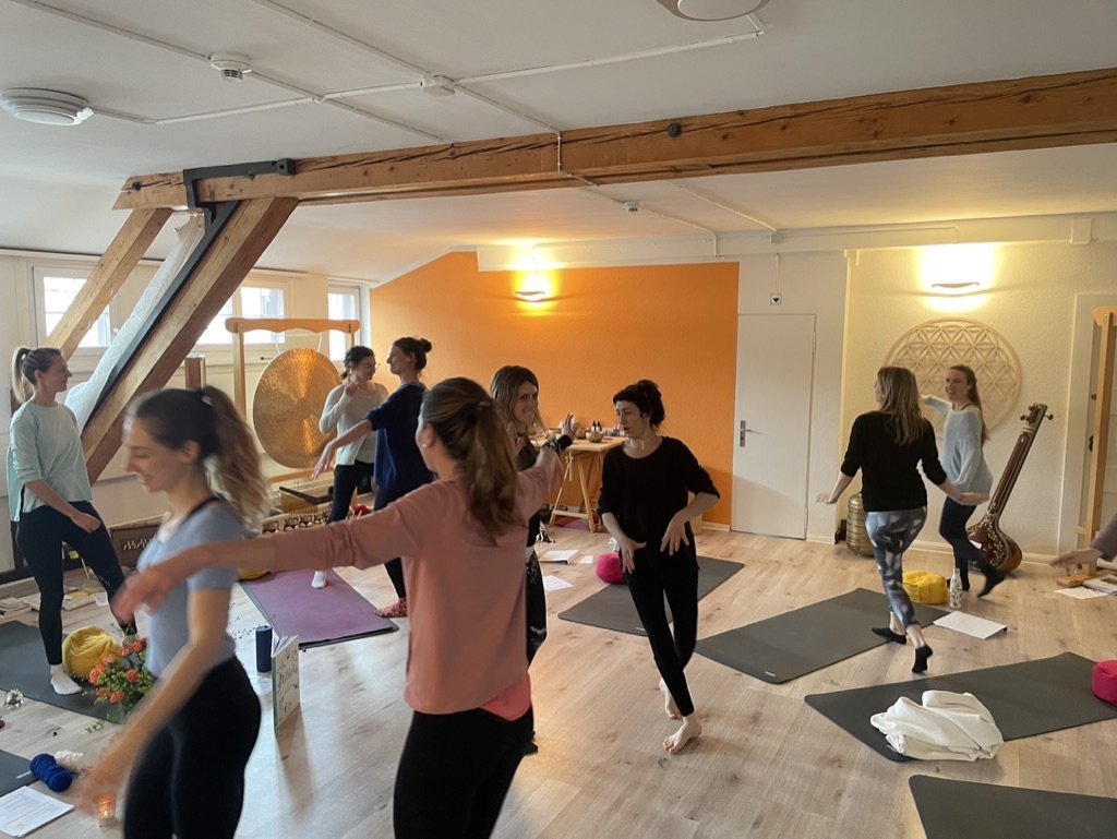 Gruppe von Frauen in einem Yoga- oder Tanzkurs in einem hell erleuchteten Raum, einige auf Yogamatten, einige stehen und unterhalten sich.