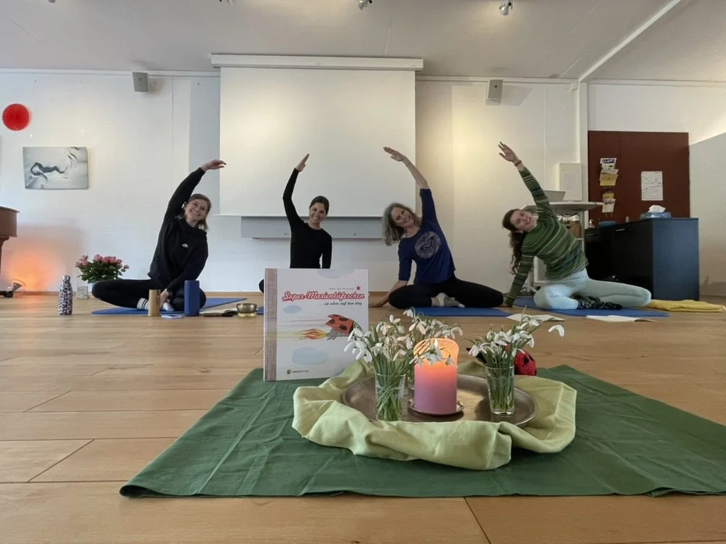 Vier Frauen in Yogahaltungen in einem Raum mit Dekoration auf dem Boden, darunter Kerzen, Blumen und ein Buch, vorne im Bild.