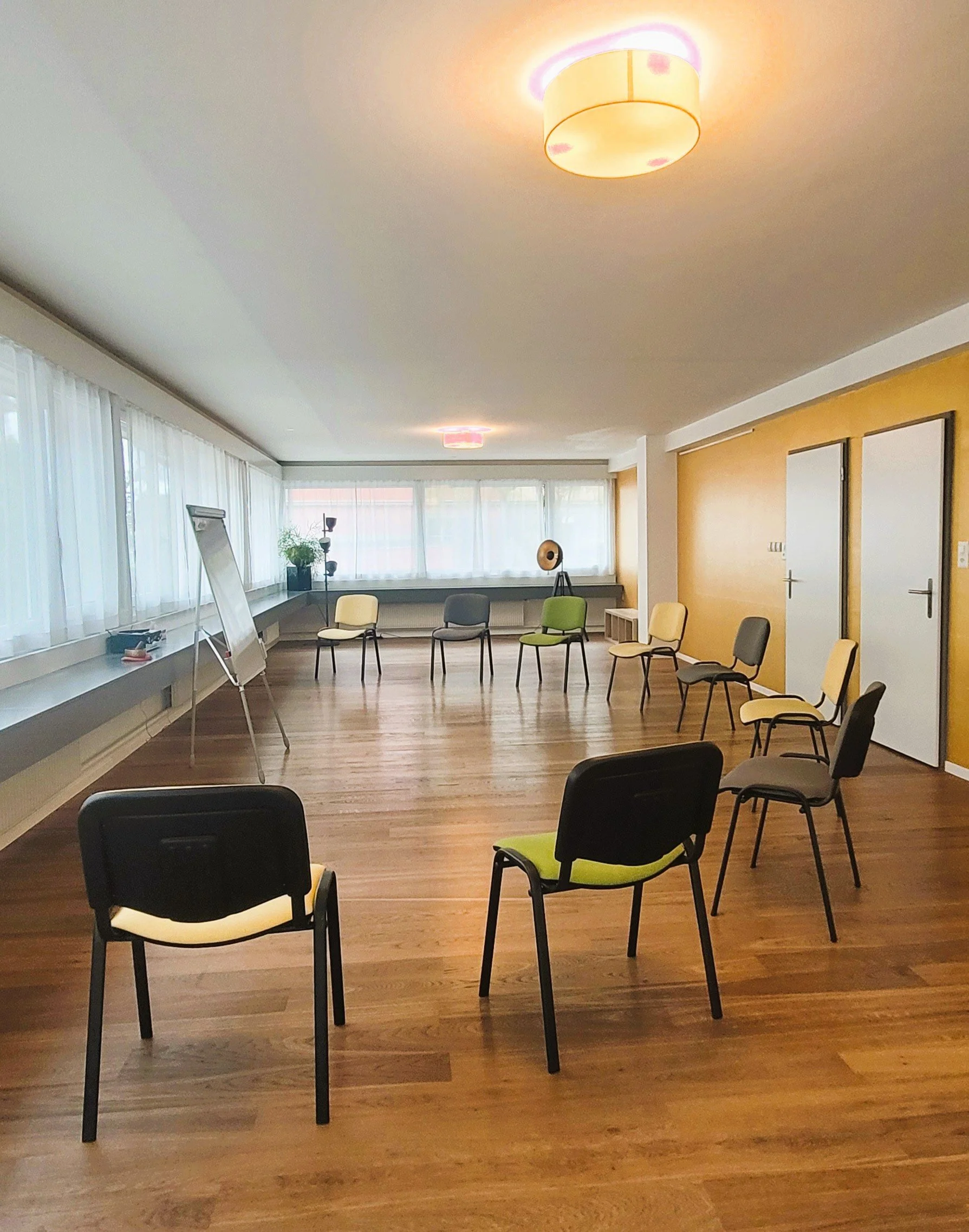Ein heller, freundlicher Seminarraum mit hellem Holzfußboden, weißen Wänden und großen Fenstern mit weißen Vorhängen. Es gibt einen Kreis aus Stühlen, einige in Schwarz, andere mit gelbem und grünem Bezug. An der Wand steht eine Flipchart. Im Hinterg