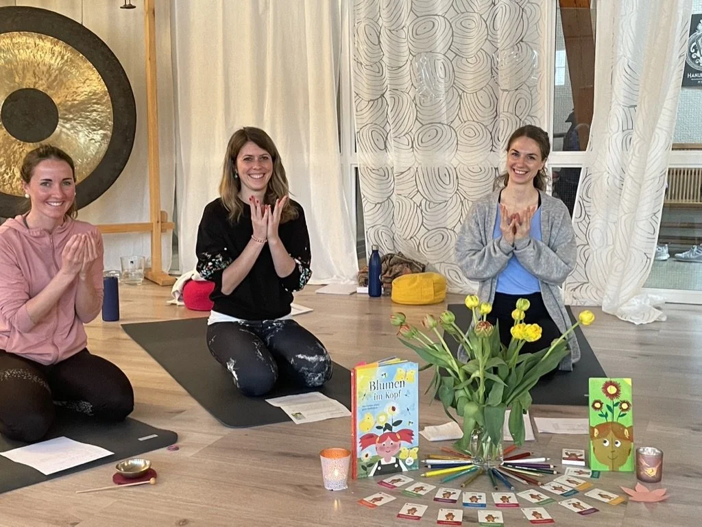 Drei Frauen beim Yoga oder einer Meditationsrunde, sitzend auf Matten im Raum, mit Blumen, Karten, und Kerzen auf dem Boden, lächelnd und in einer entspannten Atmosphäre.