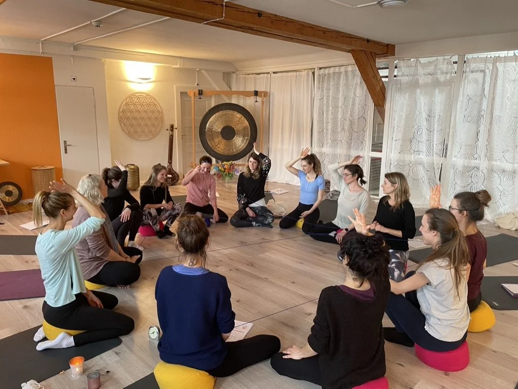 Eine Gruppe von Frauen sitzt im Kreis auf Yogamatten und bunten Sitzkissen in einem hellen Yoga-Studio, in dem sie an einer Meditations- oder Achtsamkeitsübung beteiligt sind.