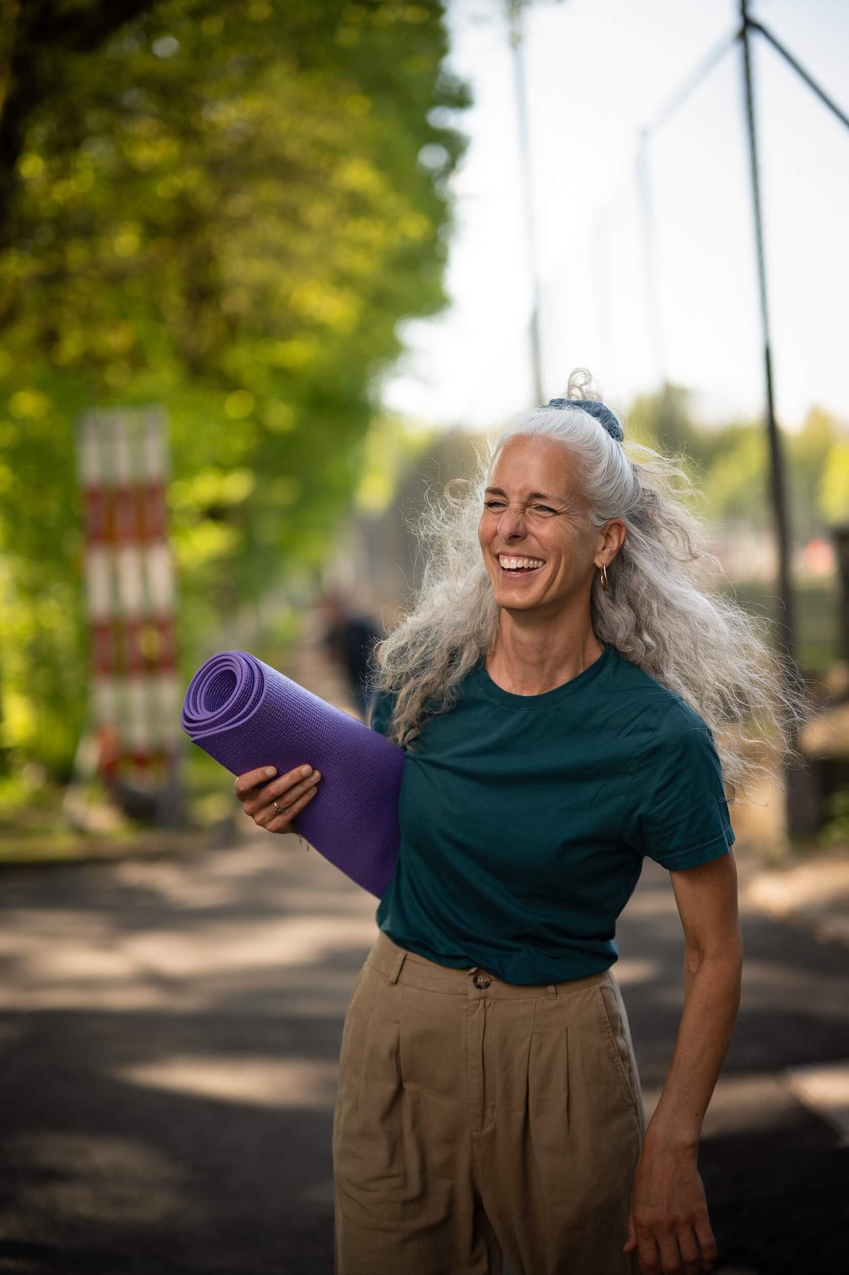Ältere Frau mit grauem, lockigem Haar, lachend, in einem Park, trägt eine dunkle T-Shirt und beige Hosen, hält eine aufgerollte Yoga-Matte in lila.