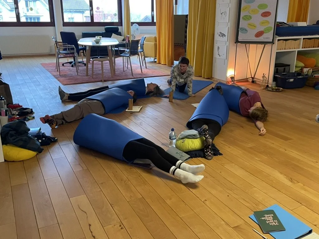 Menschen nehmen an einem Yoga- oder Entspannungsunterricht in einem Raum mit Holzboden und großen Fenstern teil, die natürliche Lichtquelle. Einige liegen auf dem Boden in Yogashapen, während eine Person sie anleitet.