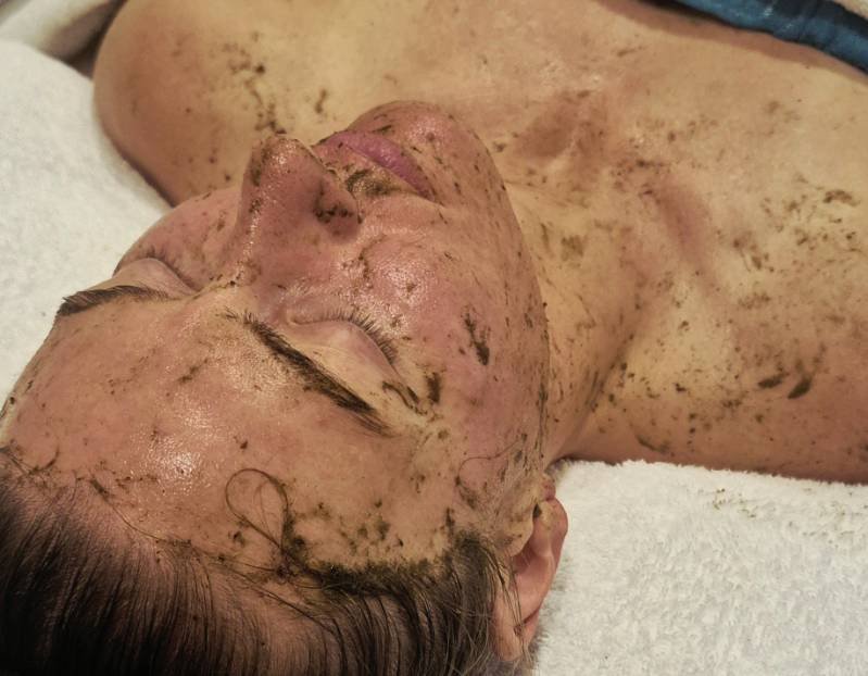 A woman receiving a facial treatment with a mud mask, lying on a spa bed with her eyes closed.