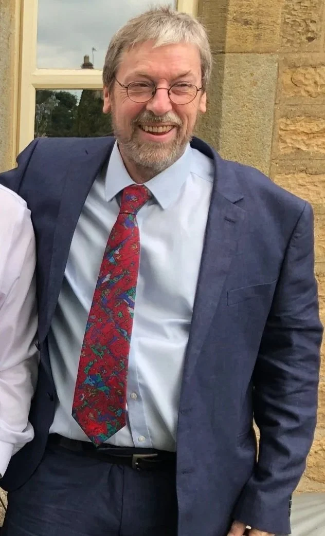 A smiling man with glasses, gray hair, and a beard, wearing a navy blue suit, white shirt, and a red patterned tie, standing outside near a brick wall and window.