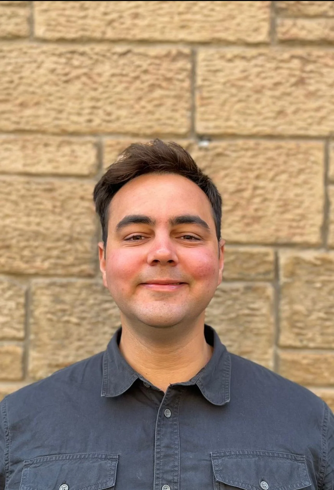 A smiling young man with dark hair wearing a black button-up shirt, standing against a beige brick wall.