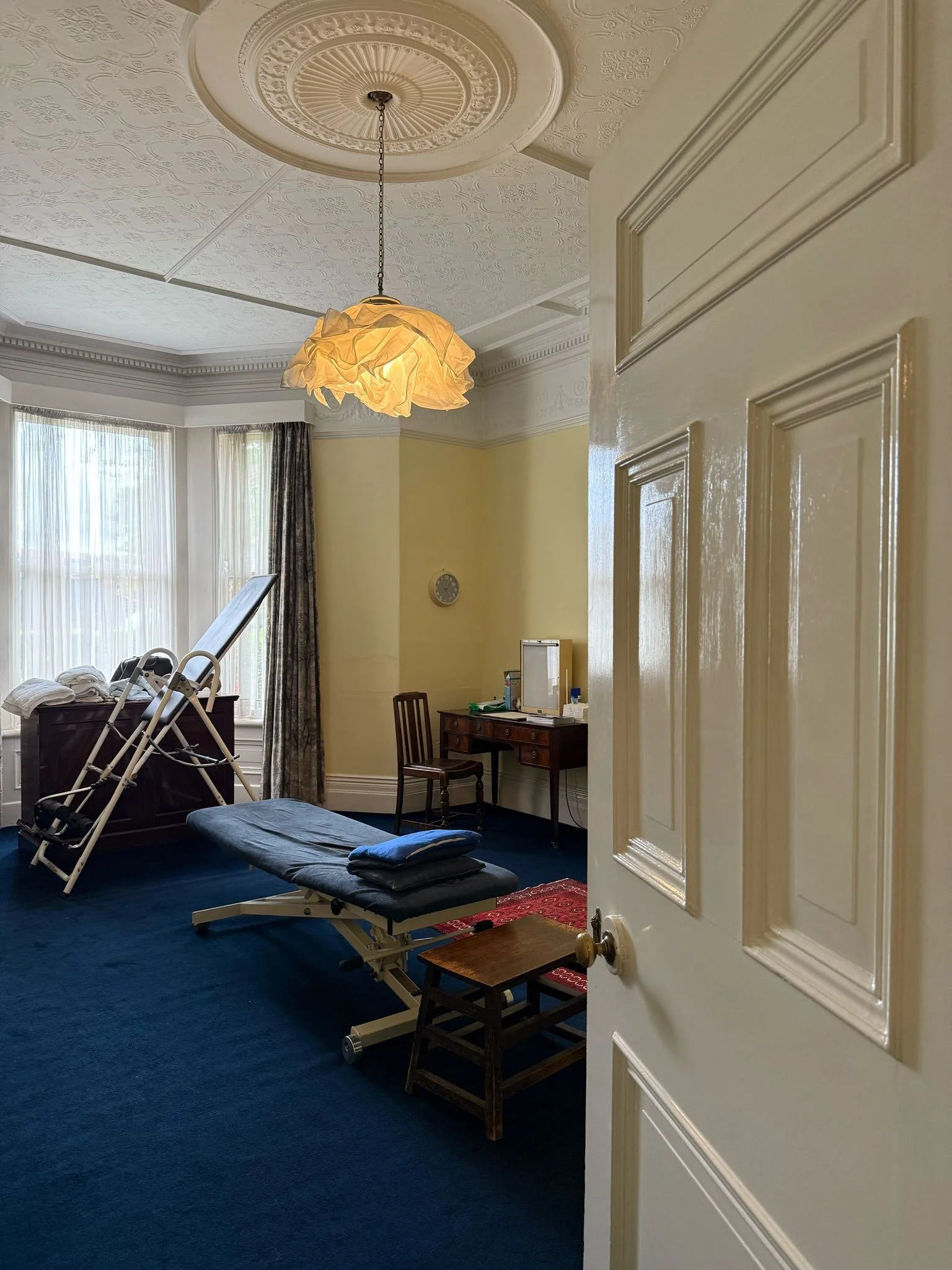 A room with a massage table, a small wooden table, and a chair, viewed from partially open door. The room has large windows with sheer curtains, patterned curtains, dark blue carpet, and a decorative ceiling with a hanging light fixture.
