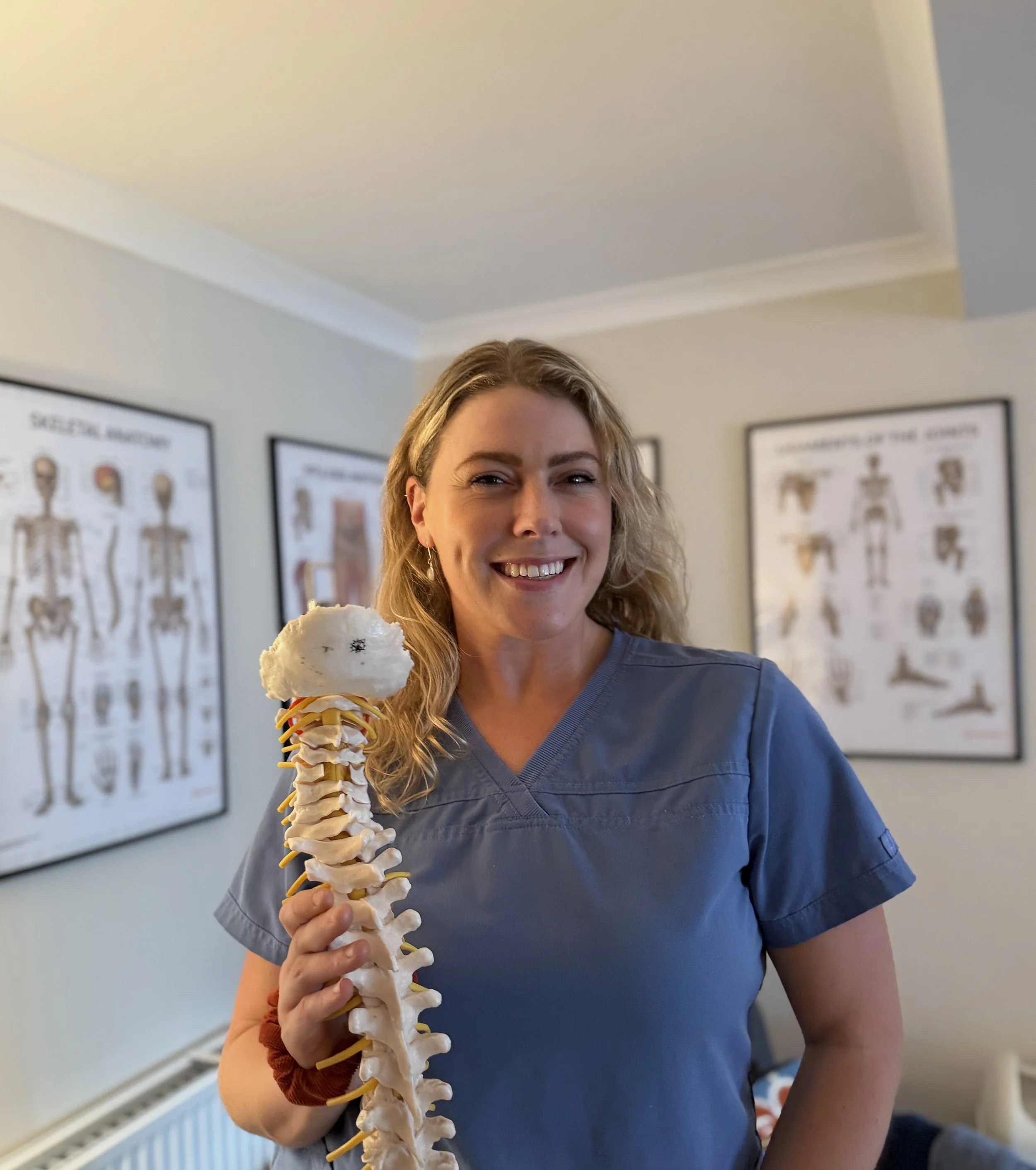 A female healthcare professional holding a model of a human spine in her hand, standing in a medical office with anatomical posters on the wall behind her.
