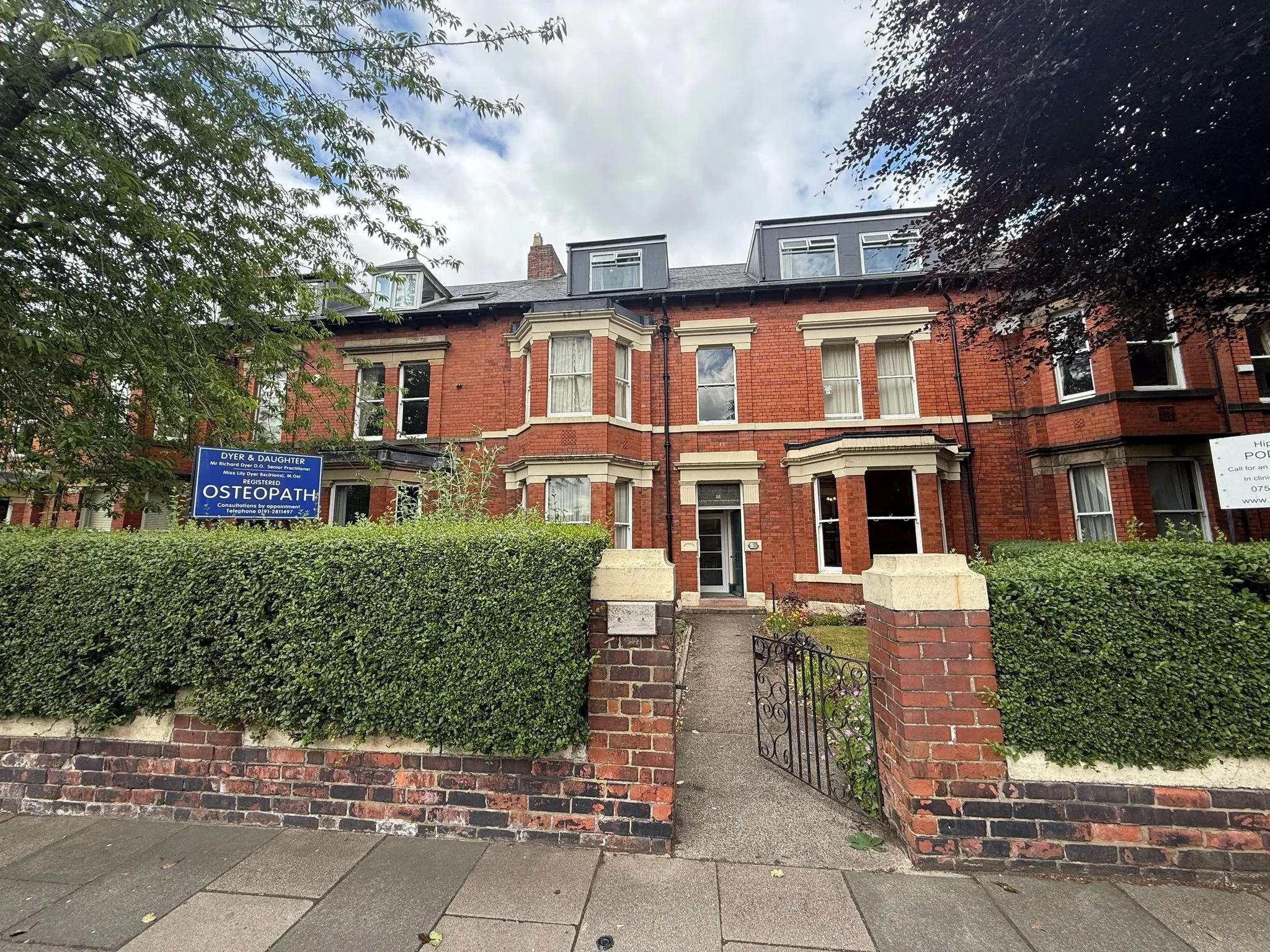 A red brick multi-story residential building with bay windows, surrounded by a green hedge and a metal gate, and a sidewalk in front.