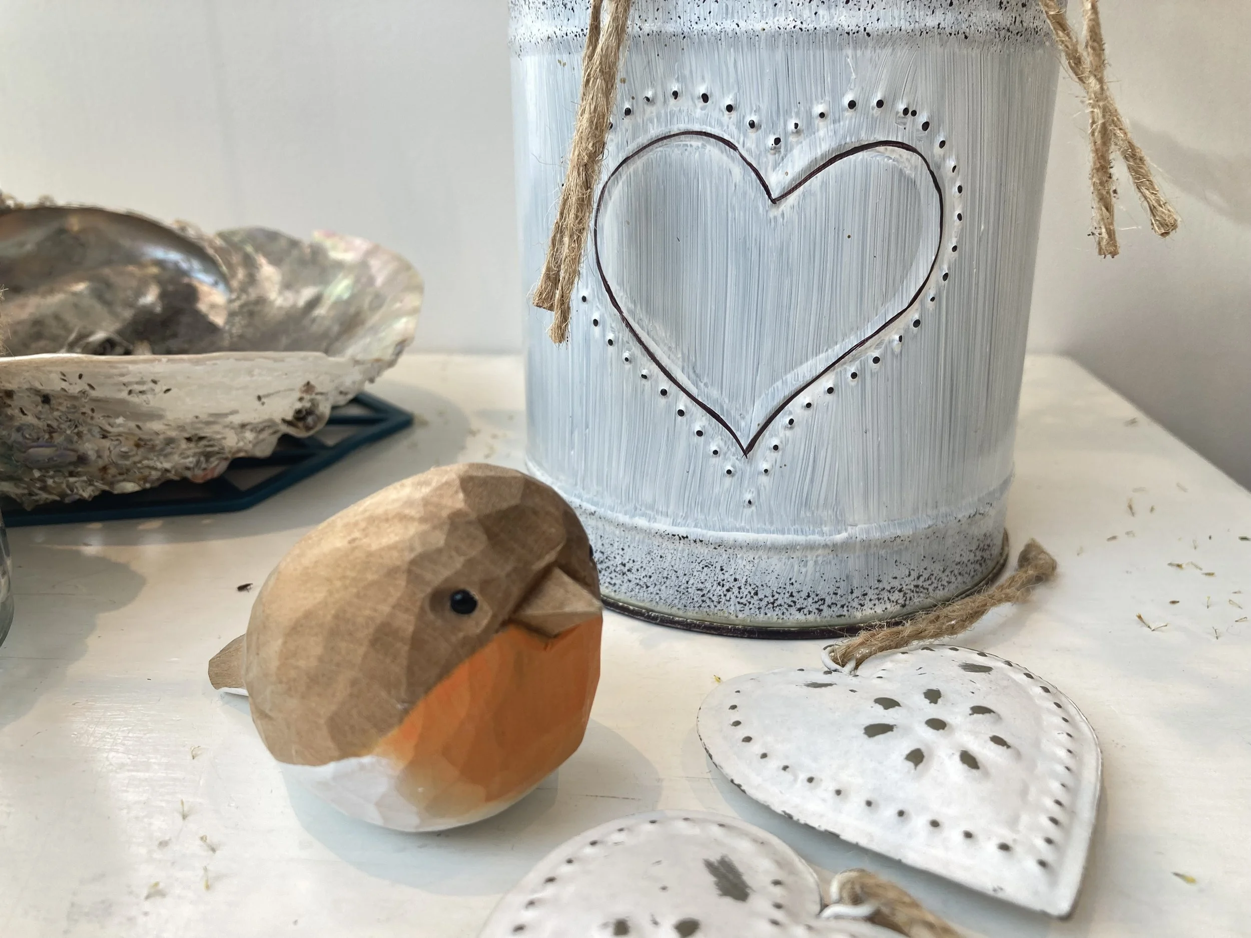 Decorative white jar with a heart and dotted outline, a wooden carved bird figurine, and white heart-shaped ornaments with dotted designs on a white surface.