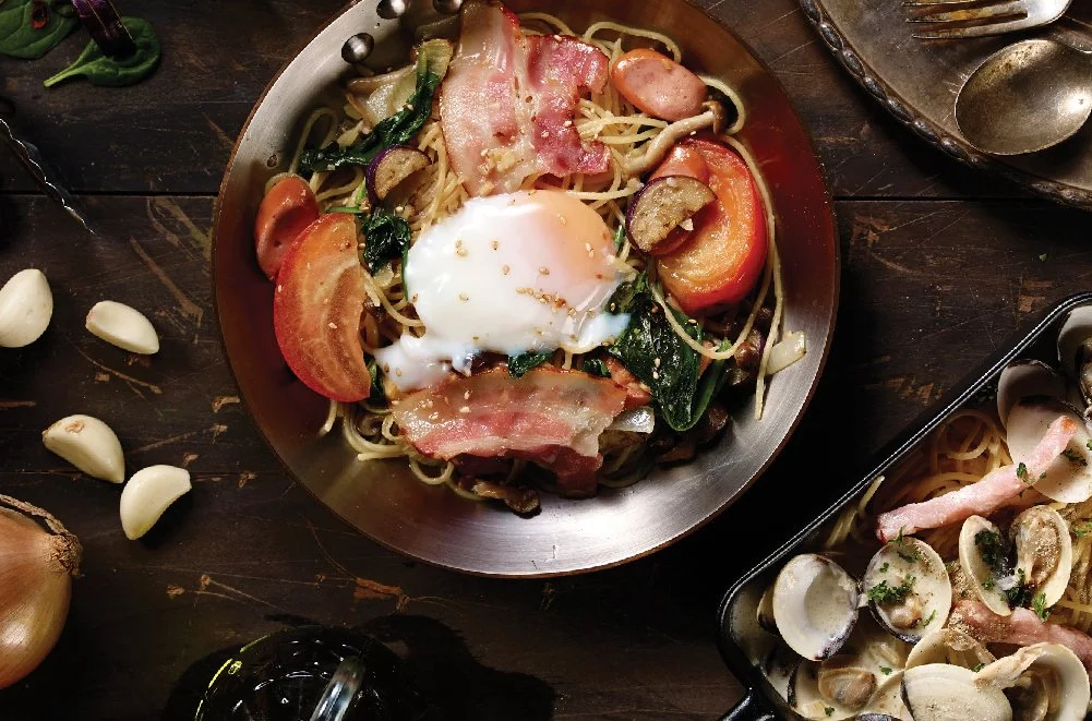 A plate of pasta with vegetables, bacon, and a poached egg, surrounded by garlic cloves, onions, and other ingredients on a dark wooden table.