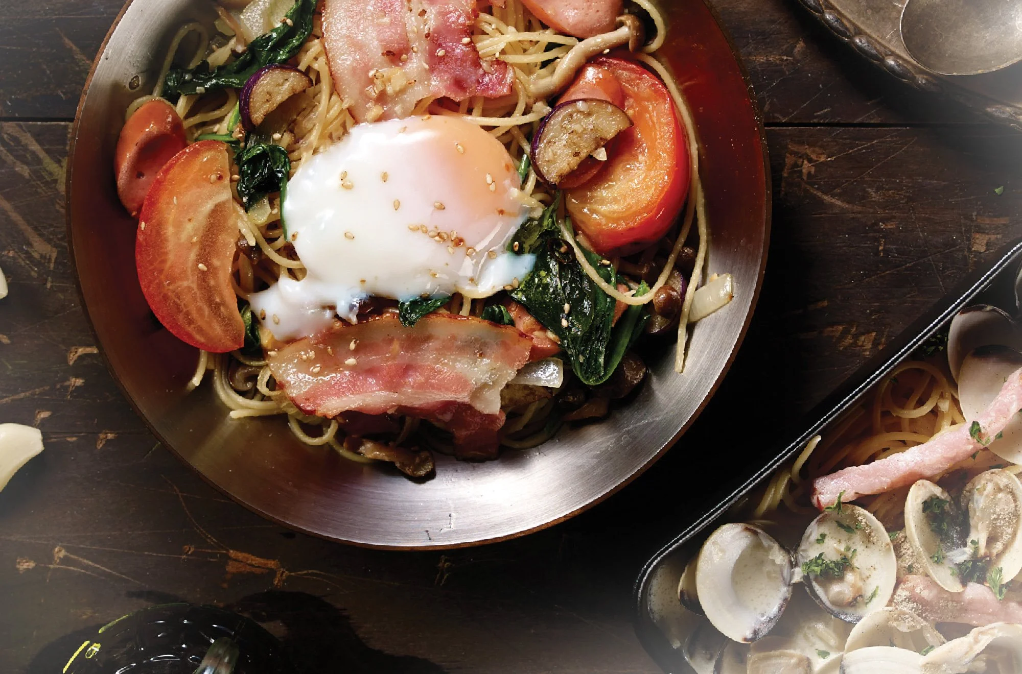A plate of pasta with vegetables, bacon, and a poached egg, surrounded by garlic cloves, onions, and other ingredients on a dark wooden table.