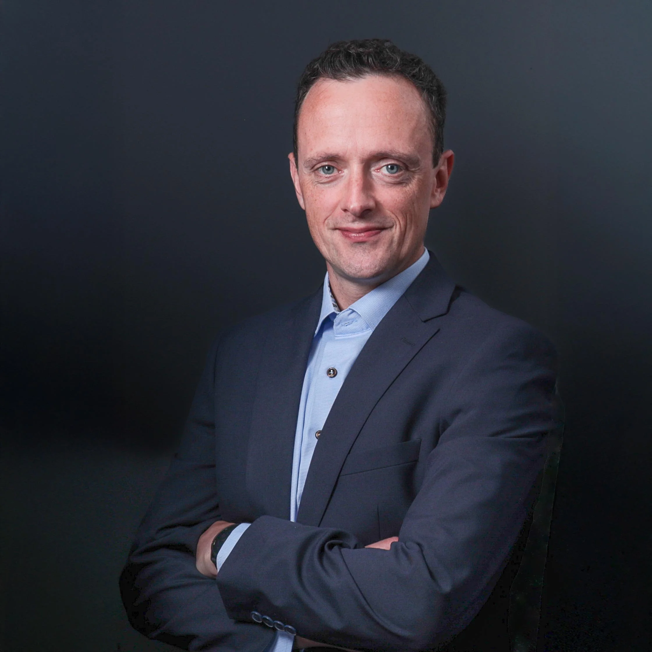 A professional man in a dark suit and light blue shirt with arms crossed against a dark background.