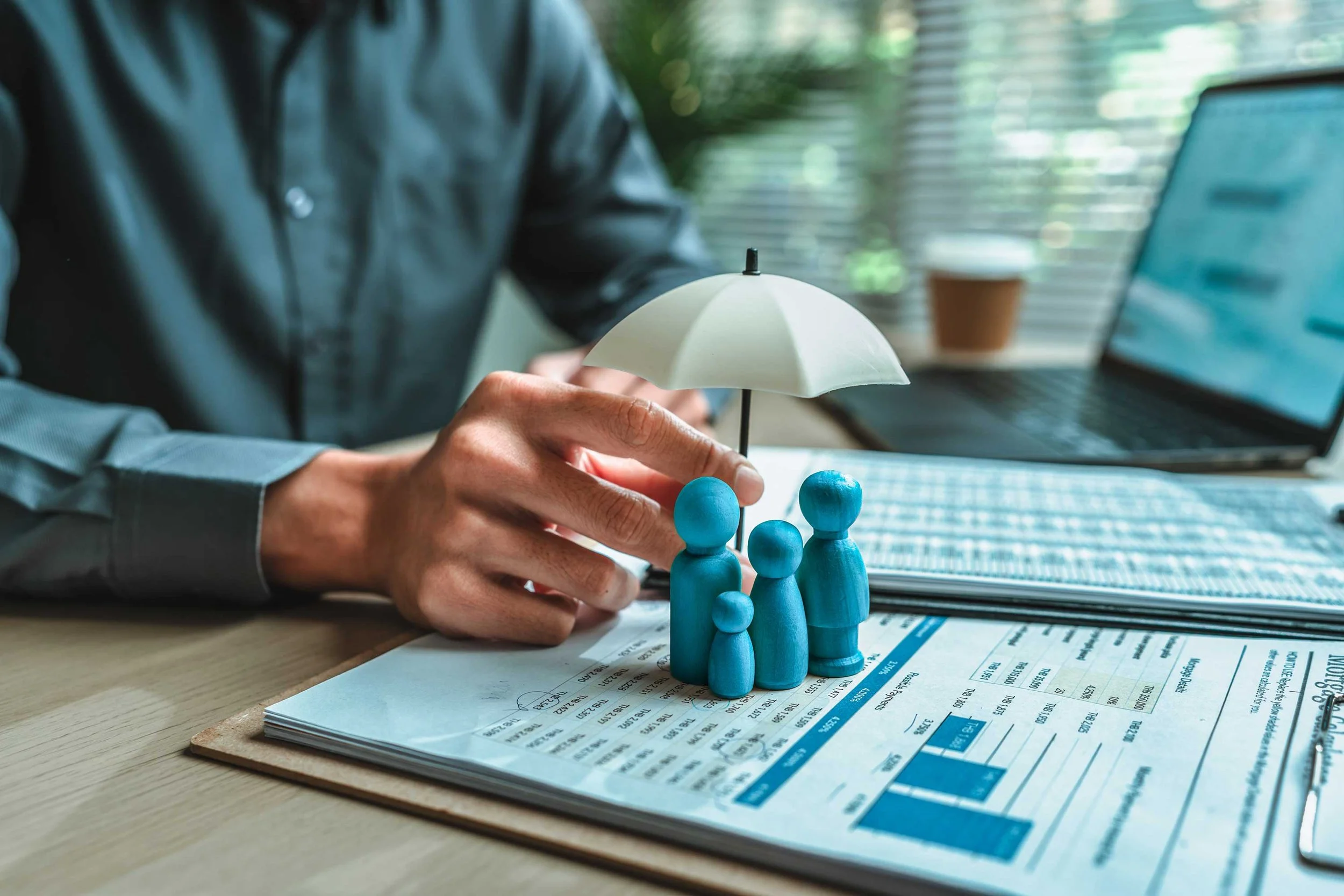 Person placing blue wooden figures under a small white umbrella on financial documents at a desk with a laptop and potted plant in background.