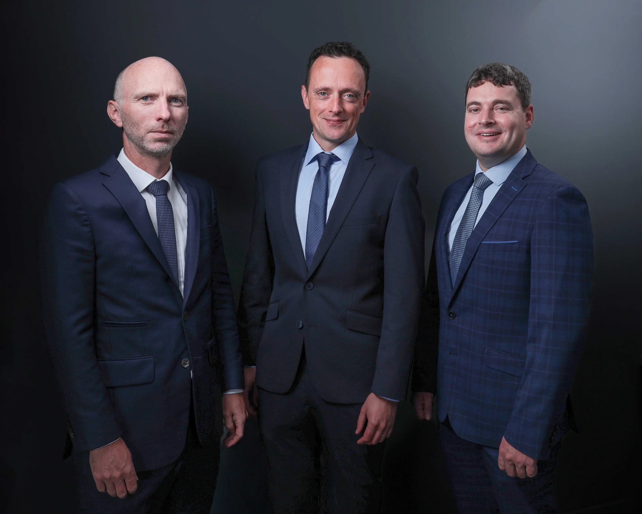 Three men in formal suits standing against a dark background.