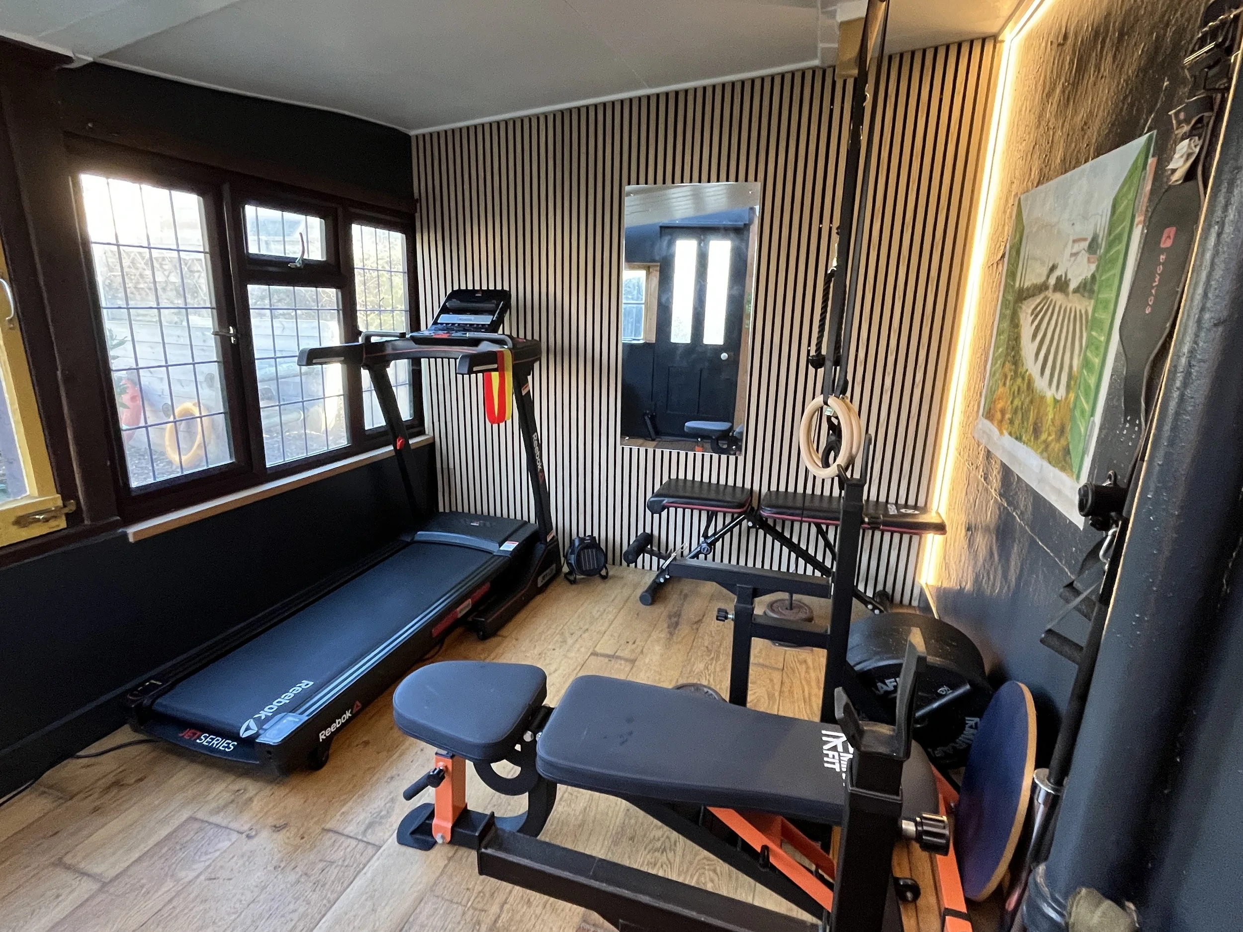Home gym with treadmill, adjustable weight bench, yoga rings, and a poster on the black wall.