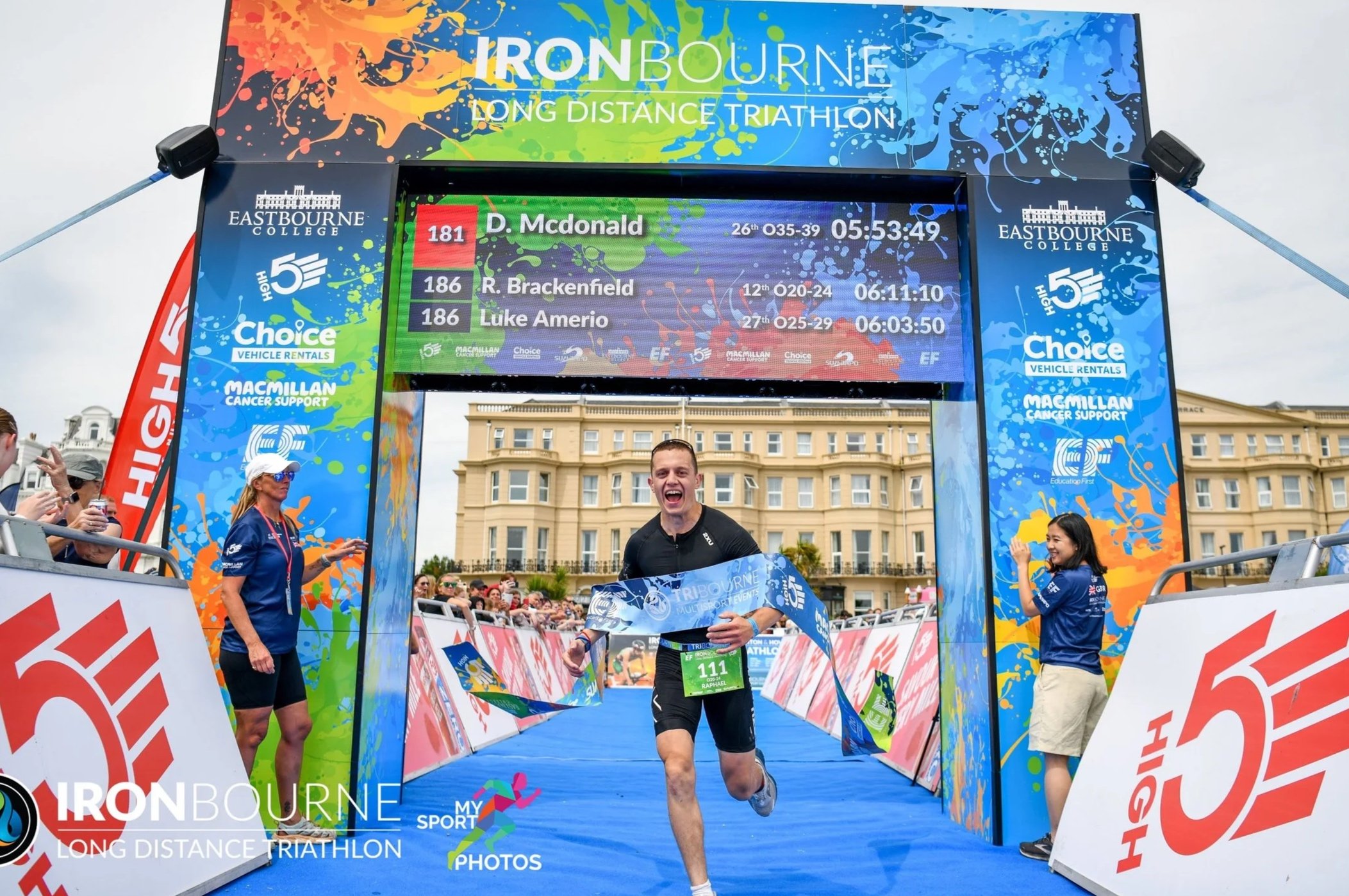 Triathlete crossing finish line at Ironbourne Long Distance Triathlon, smiling and holding a race banner, with spectators and event staff around, digital scoreboard above displaying race times and participant names.
