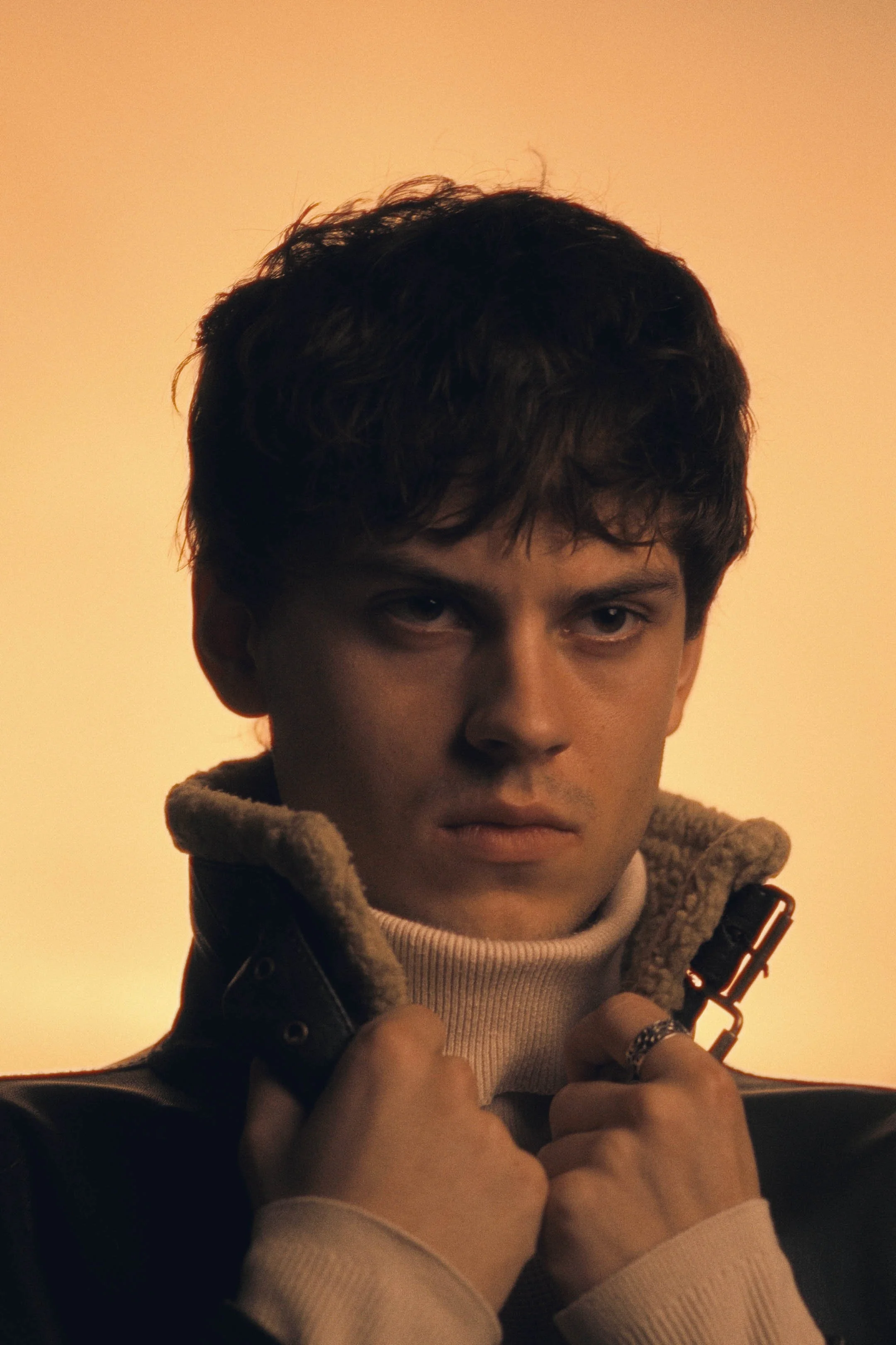 Fashion portræt af ung mand med mørkt hår og stålfast blik, i læderjakke med lammeskindsfor, på lysende orange baggrund - Studiesetting