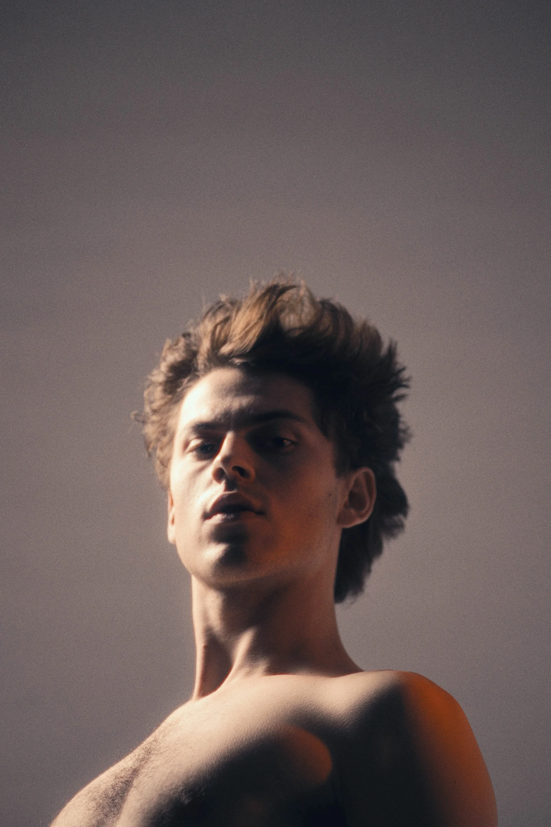 Fashion portræt af ung mand med mørkt hår og stålfast blik, i bar overkrop i kontrastfyld lyssætning, næsten skulpturel på grå baggrund - Studiesetting