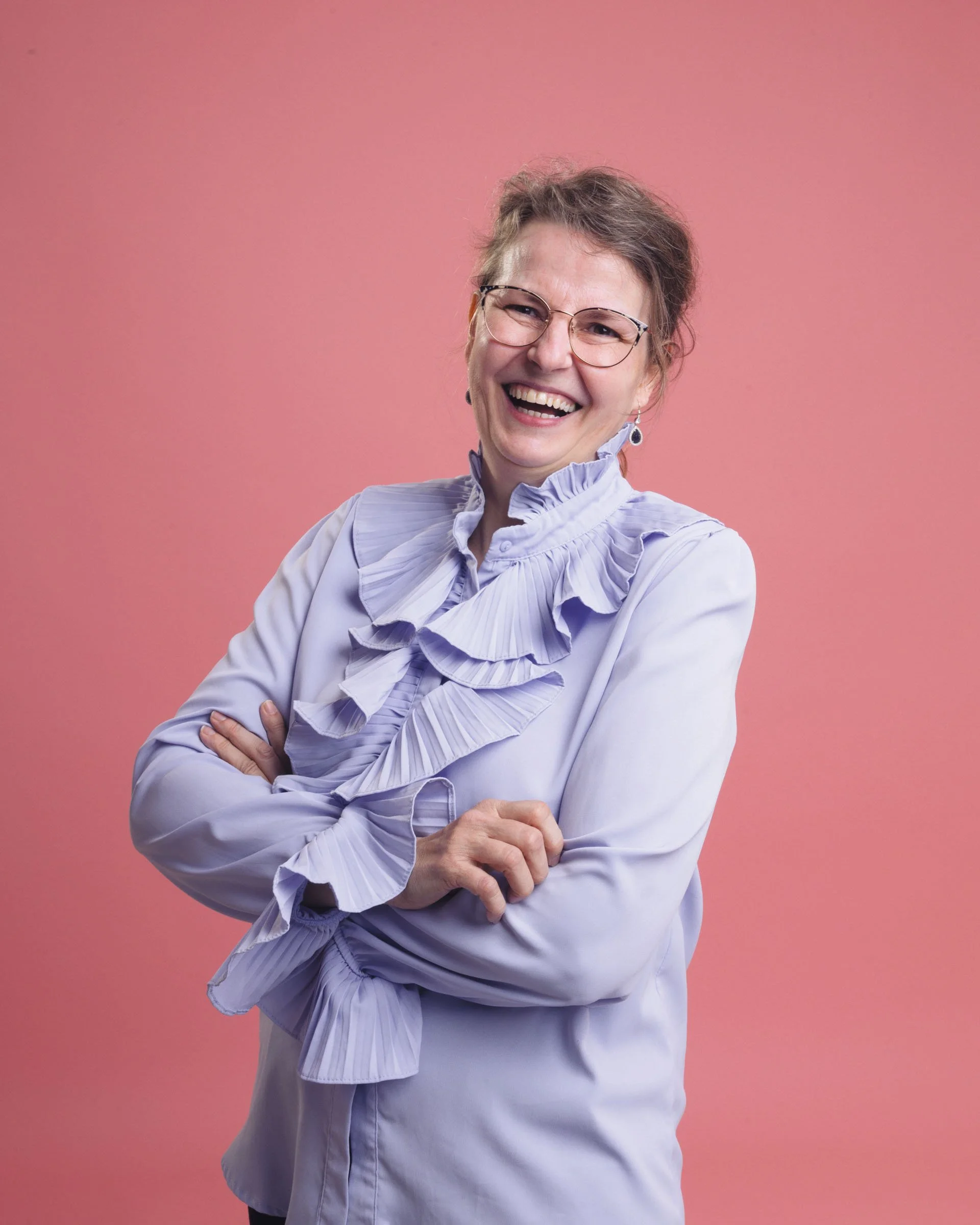 Farverigt portræt af leende kvinde i lilla skjorte, på lyserød baggrund.