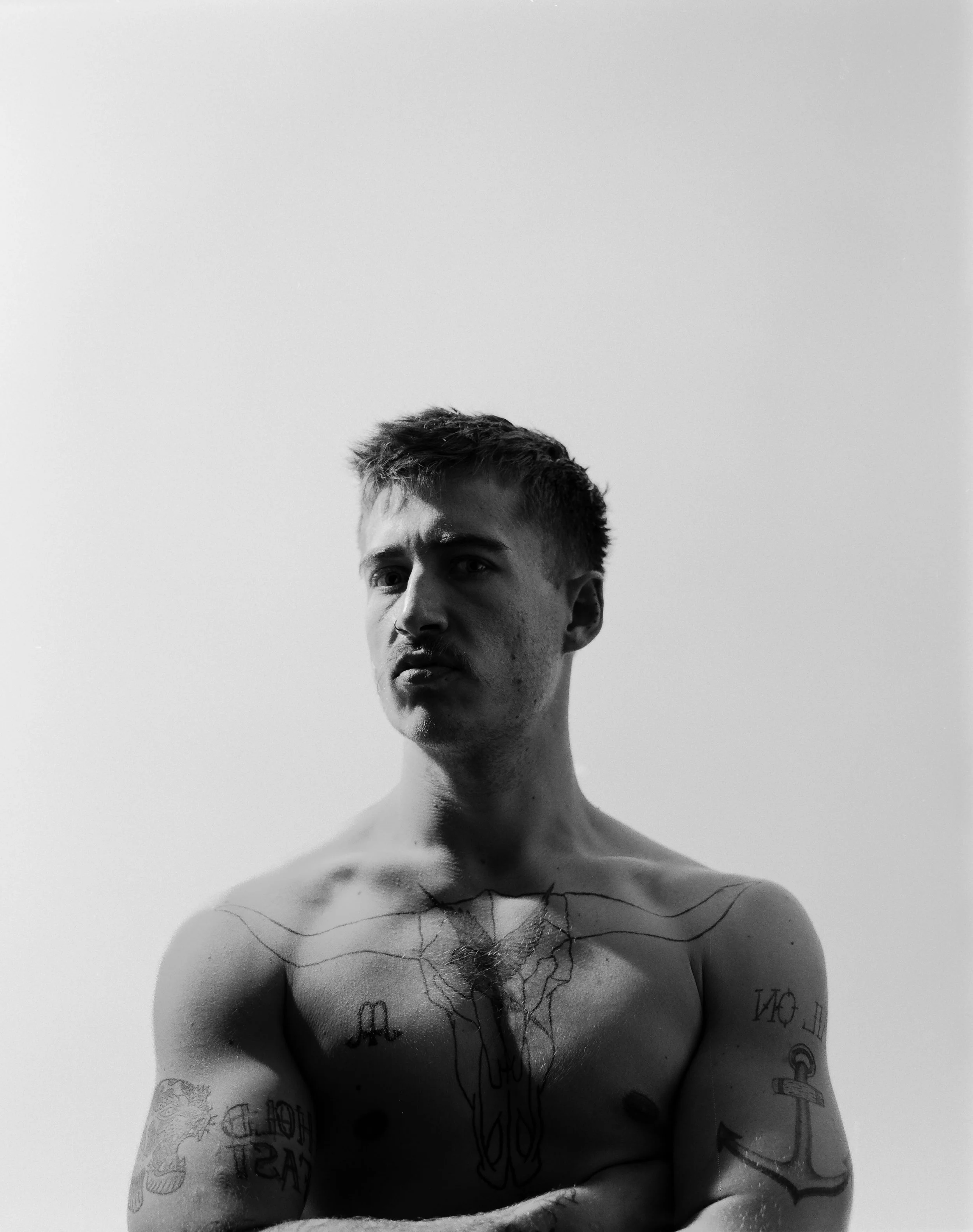 Personligt portræt af ung mand i bar overkrop, med stolt blik og tatoveringer, i kontrastfyldt lys - Studiesetting