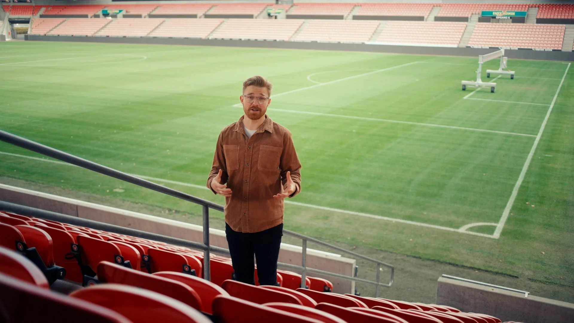 Stillgrab fra produktionen hvor Buster står på tribunerne ved MCH Arena. Han har den grønne fodboldbane i ryggen. Der er mange ledende linjer.
