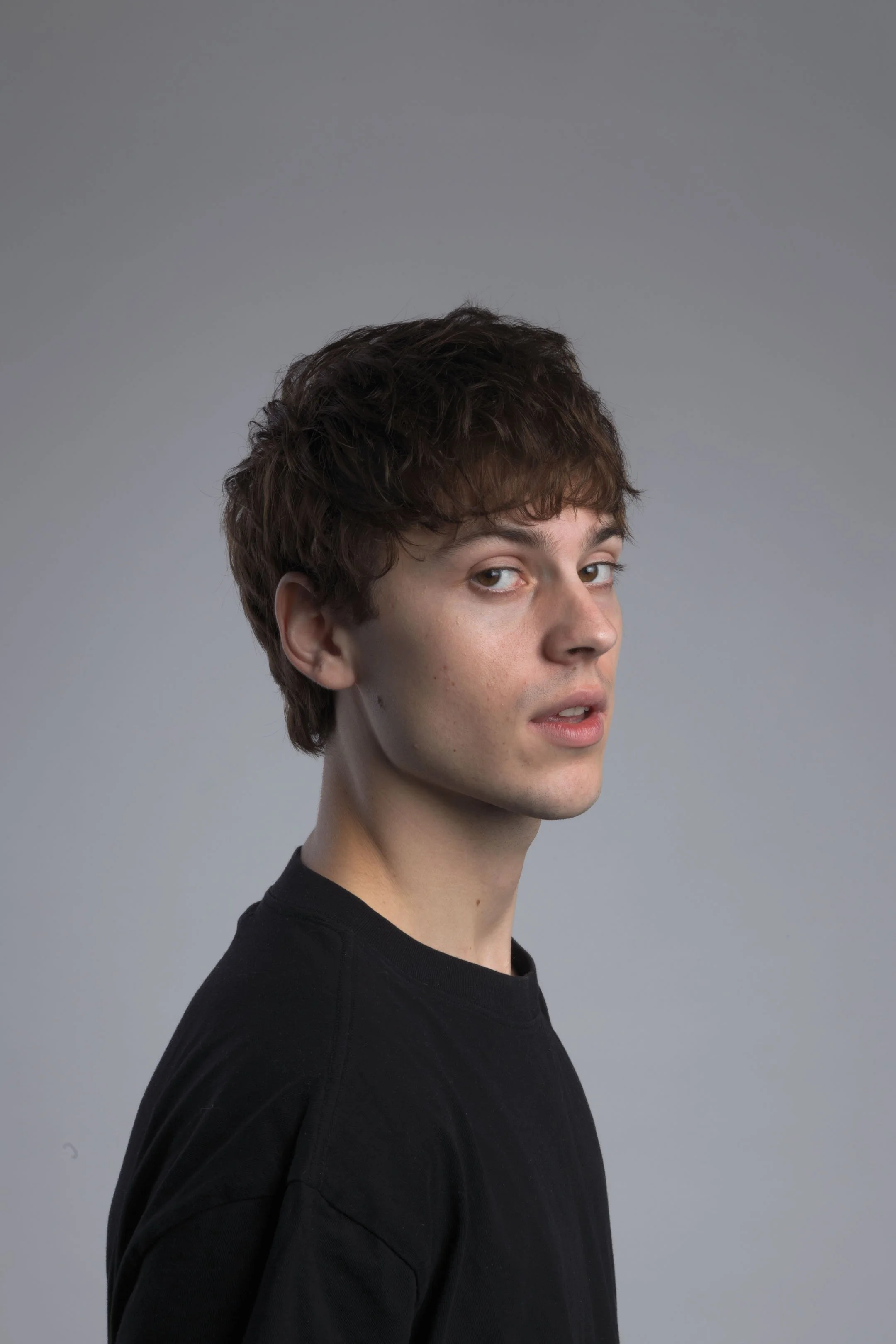 Portræt af ung mand i profil med mørkt hår med et glimt i øjet, i sort t-shirt på grå baggrund - Studiesetting