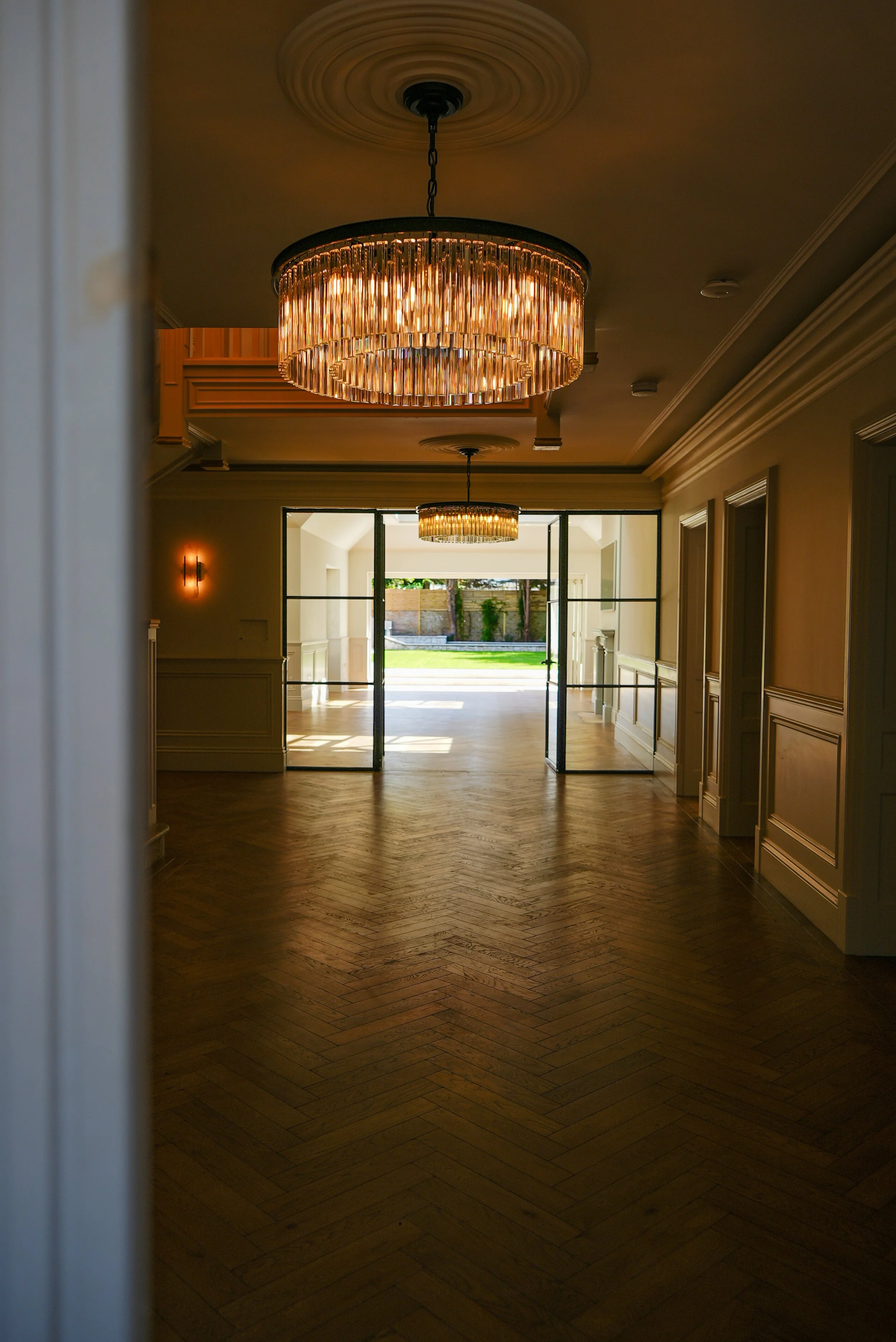 Interior of a luxurious room with two elegant chandeliers, wooden parquet flooring, and open glass doors leading to a bright outdoor area.