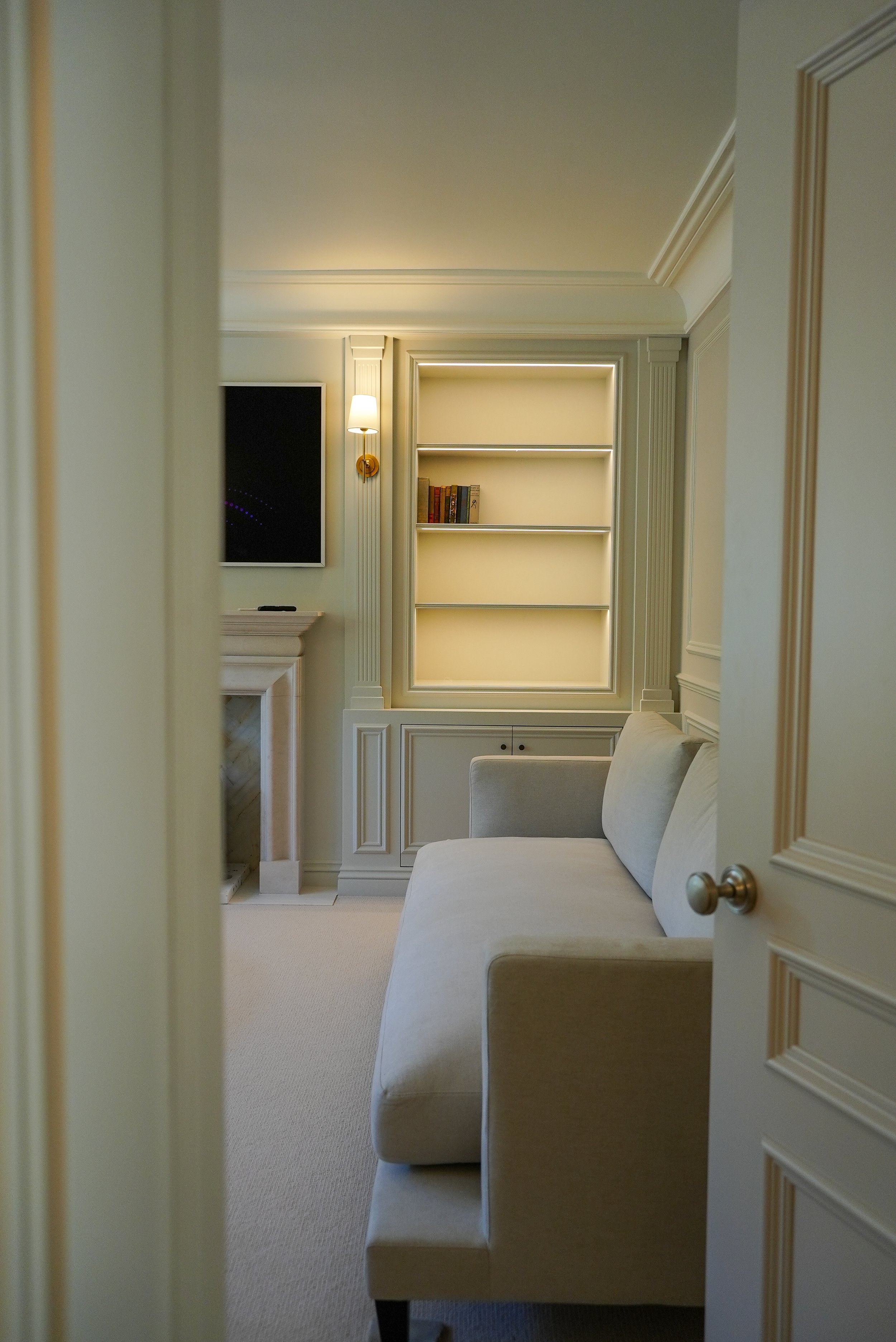 View through a partially open door into an elegant, well-lit living room with a cream-colored sofa, built-in bookshelves, a fireplace, a wall-mounted television, and a wall sconce.