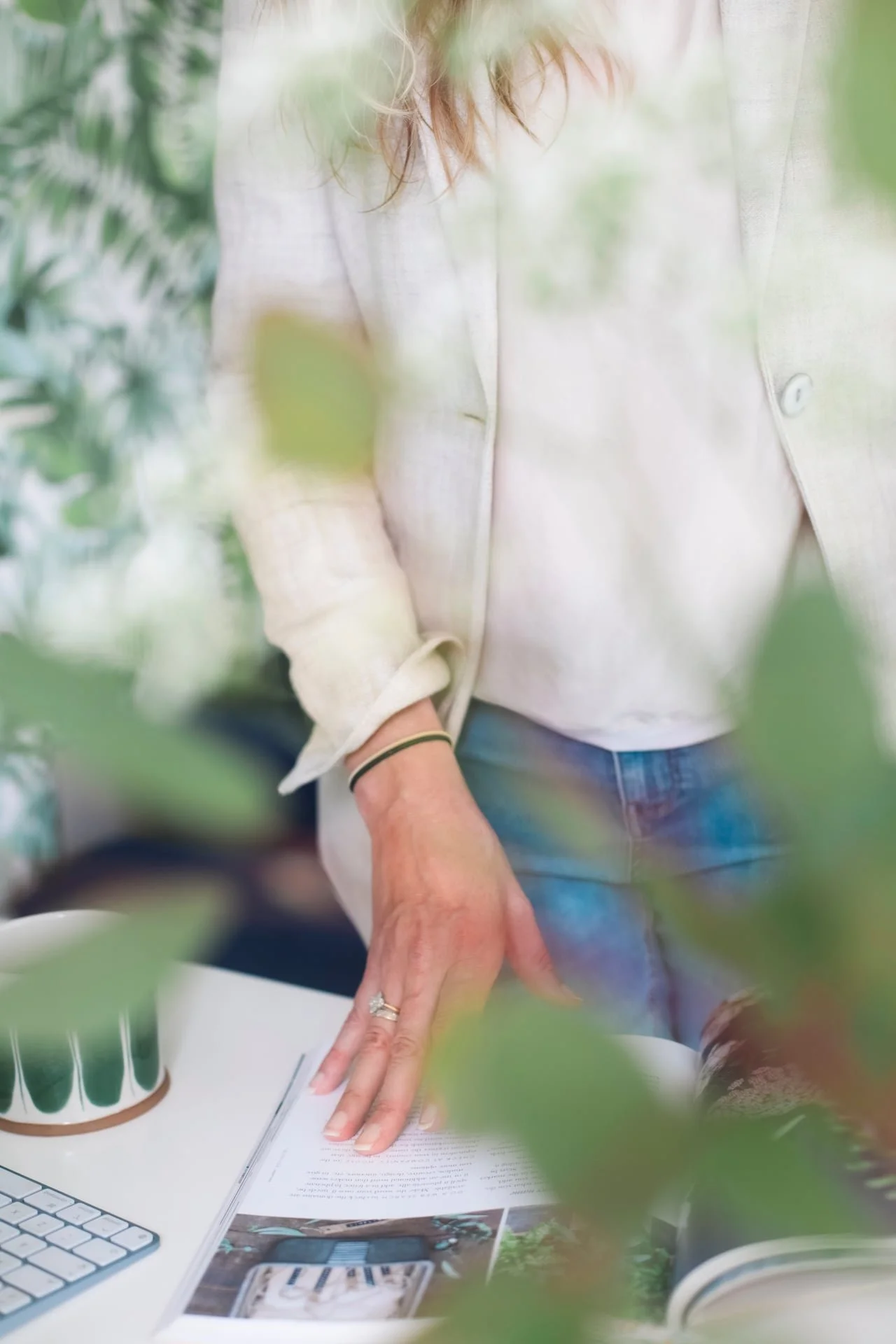 Person wearing a light-colored plaid shirt, standing at a white desk with a computer and paper, surrounded by green leaves in the foreground.