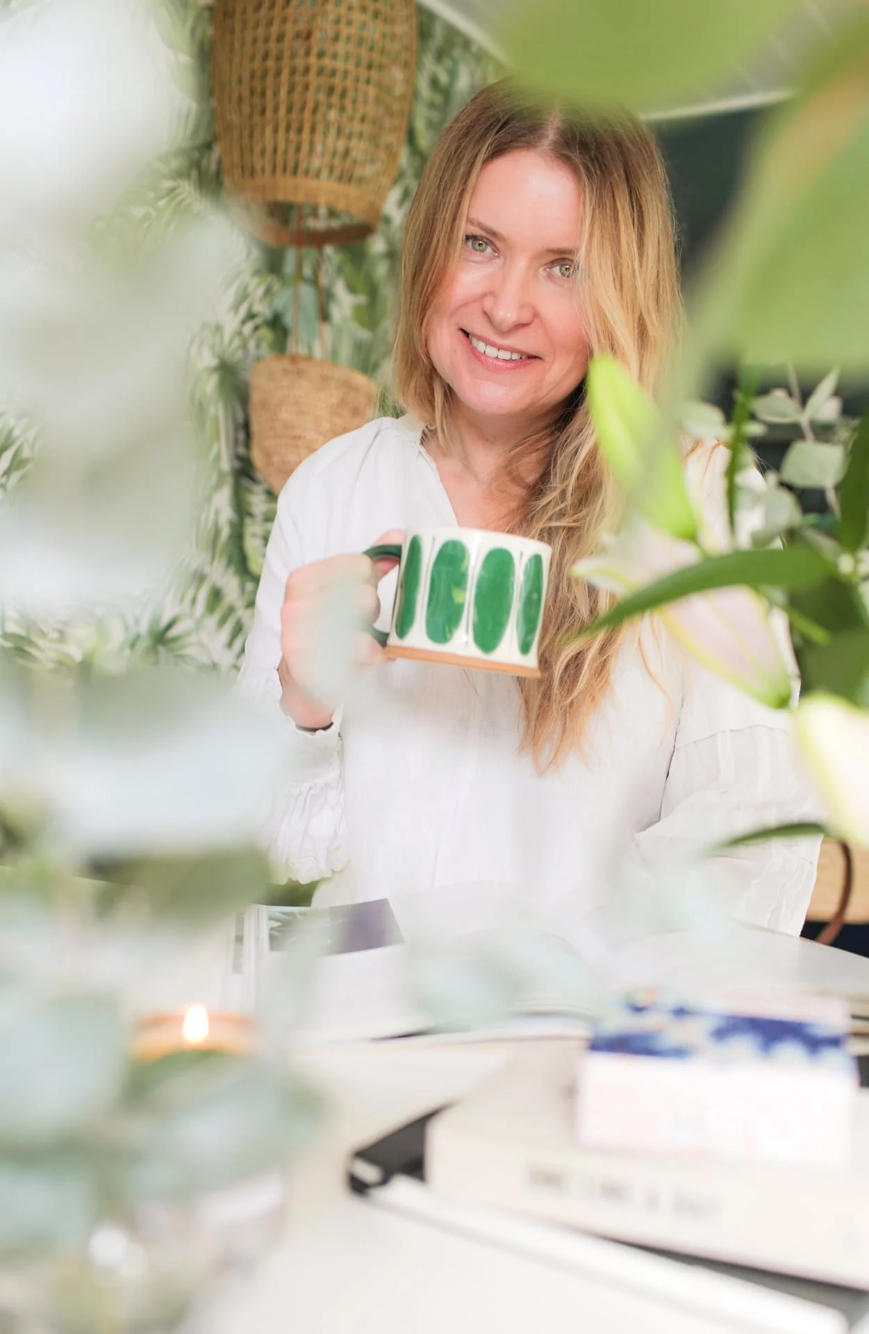 A woman with blonde hair smiling and holding a green and white mug, surrounded by plants indoors.