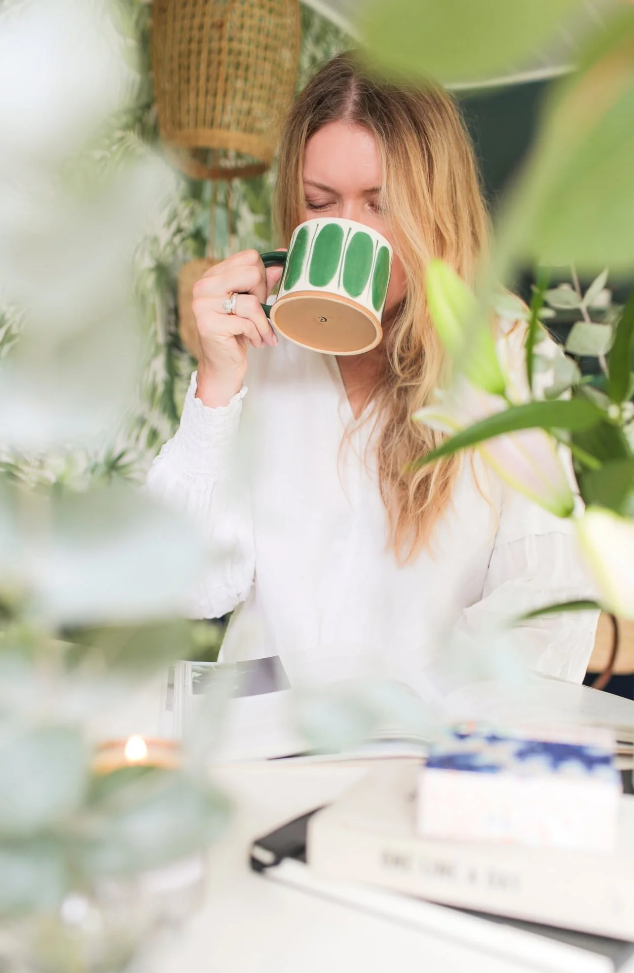 A woman with long, wavy, blonde hair drinks from a white mug with green vertical stripes while sitting at a table surrounded by green plants.