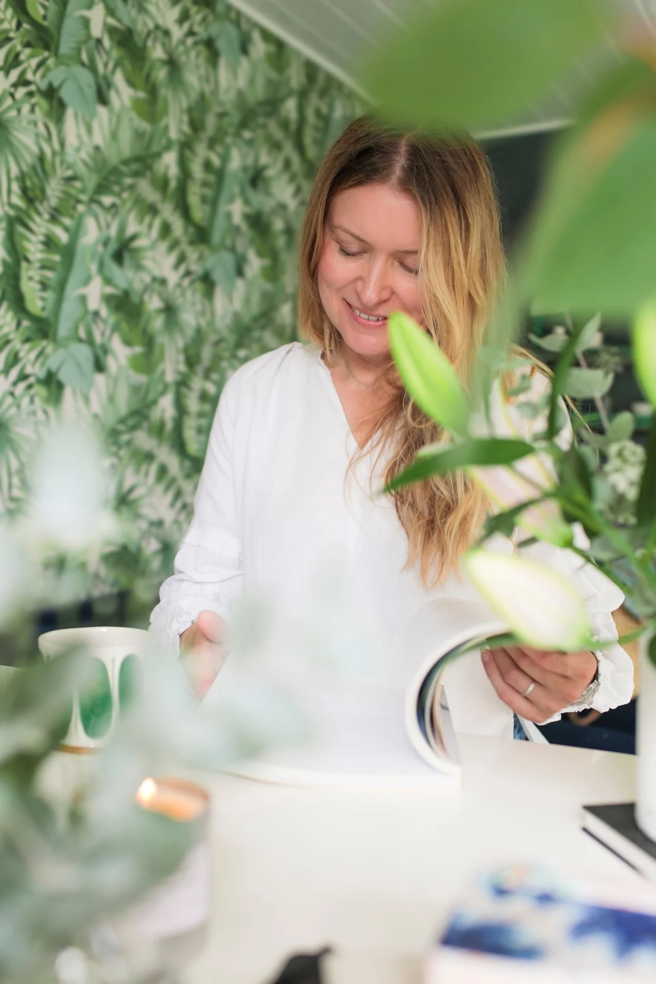 A woman with long, wavy blonde hair reading a book while sitting at a white table surrounded by green plants in a bright room.
