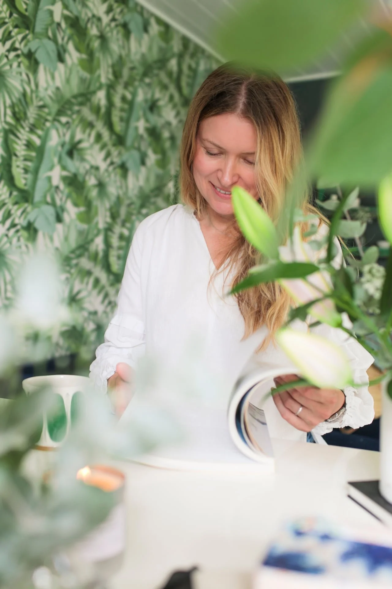 A woman with long, wavy hair wearing a white shirt looking at a magazine in a greenery-filled environment.