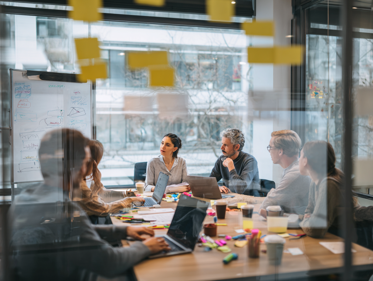 A diverse group of people in a business meeting room, engaged in discussion, with laptops, notebooks, and coffee cups on the table, viewed through a glass window with yellow sticky notes attached.