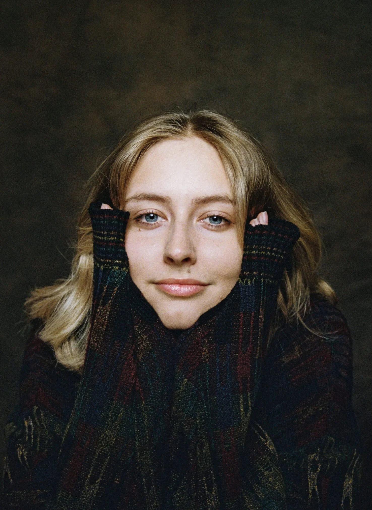 Close-up of a woman with blonde hair and blue eyes, wearing a dark plaid sweater, with hands behind her head against a dark background.