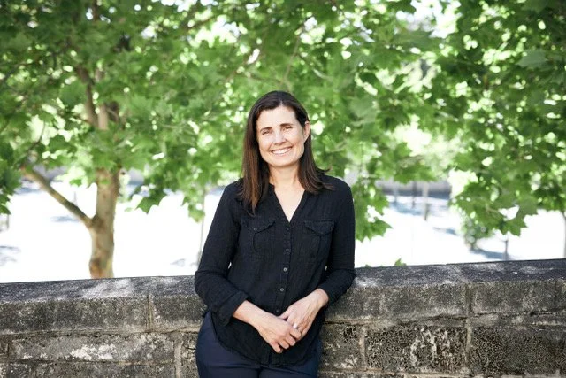 A woman with dark brown hair wearing a black shirt, smiling and standing outdoors in front of green trees and a stone wall.