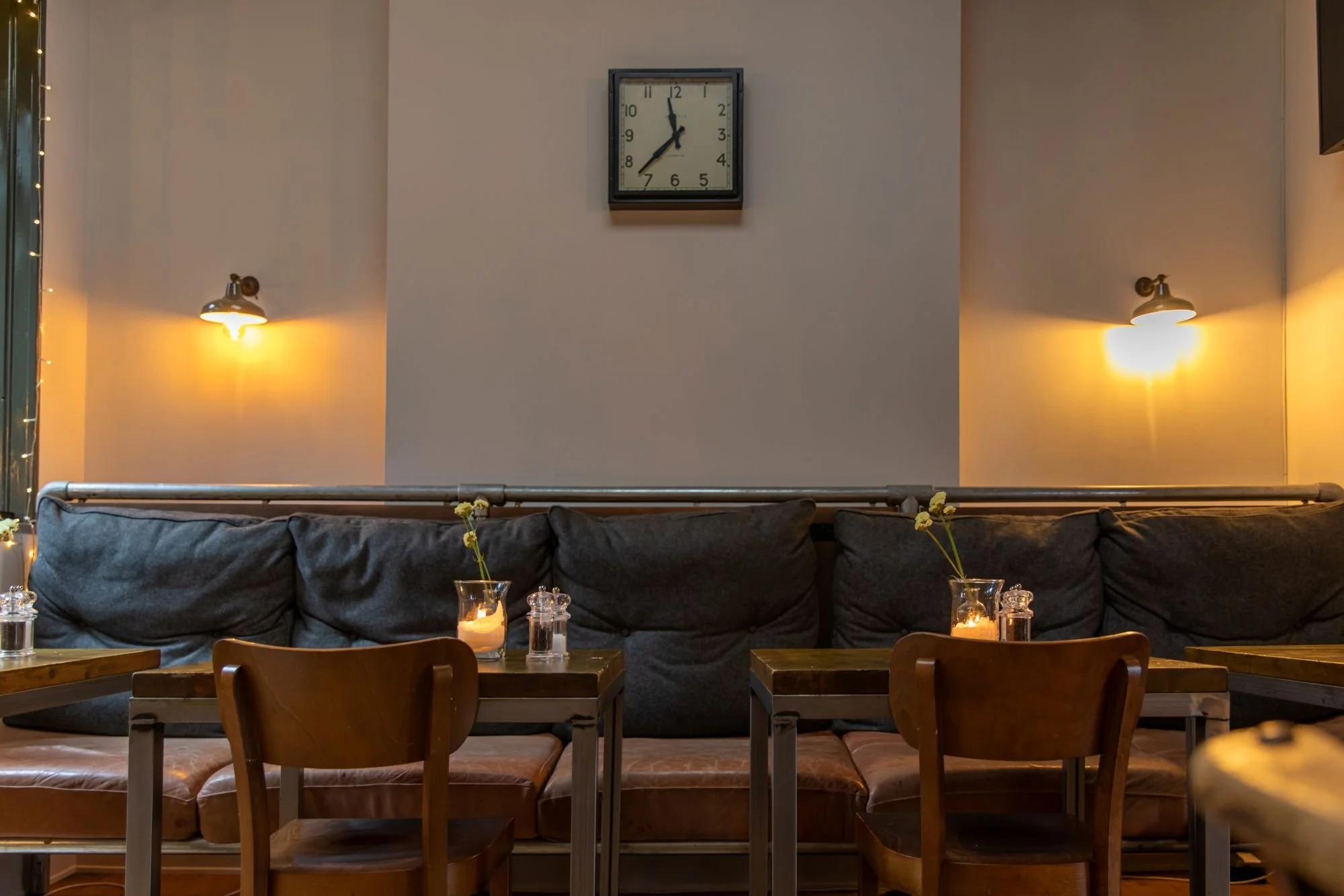 Cozy restaurant interior with tables, chairs, candles, and wall-mounted lights, featuring a clock on the wall.