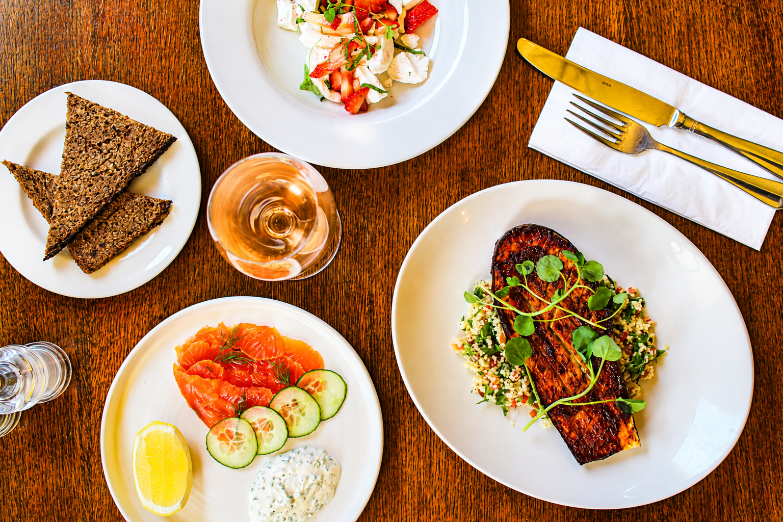 A dining table featuring a glass of rosé wine, a plate of grilled salmon with greens and couscous, a bowl of strawberry and mozzarella salad, a plate with smoked salmon, cucumber slices, lemon wedge, and a dollop of sauce, and a small plate of crisp bread triangles.