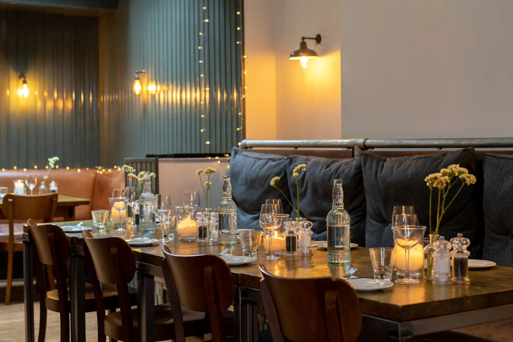 A cozy restaurant interior with a long wooden table set with glass bottles, candle holders, wine glasses, plates, and flowers. Warm lighting and fairy lights create an inviting atmosphere.
