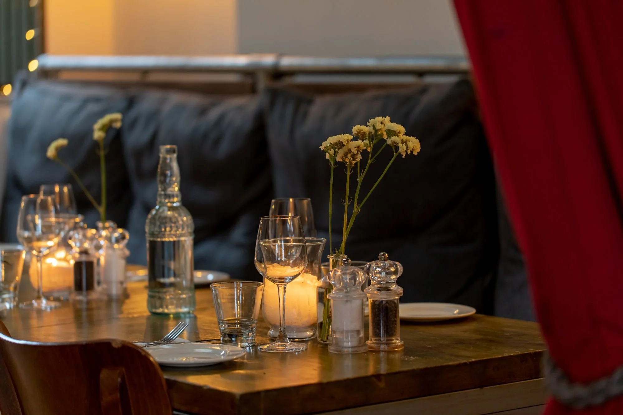 A warmly lit dining table set with white plates, wine glasses, water glasses, carafes, salt and pepper shakers, a candle, and vases with white flowers, with dark cushions and a candlelit background.