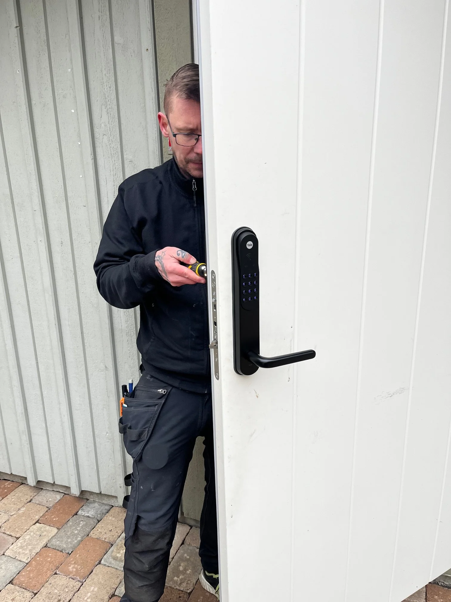 En man med glasögon och arbetskläder som installerar en elektronisk låslock på en vit dörr utomhus.