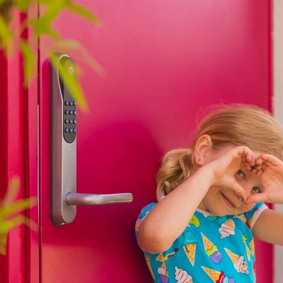 En flicka med lockigt hår i en blå tröja med isglass och koner, leende och gör en hjärtformad gest med händerna framför ansiktet, framför en rosa dörr med ett lås.